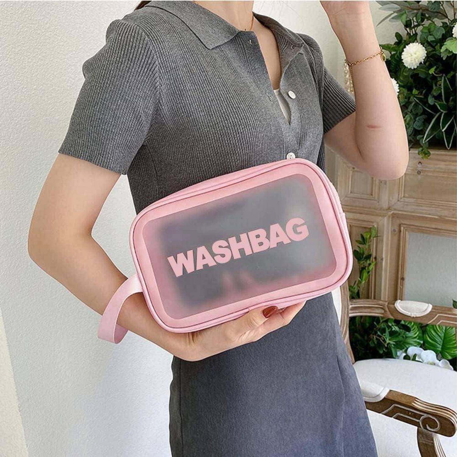 Person hält eine rosa Kulturtasche mit dem Wort ‚WASHBAG', der in einem Raum mit Pflanzen und einem Stuhl im Hintergrund steht.
