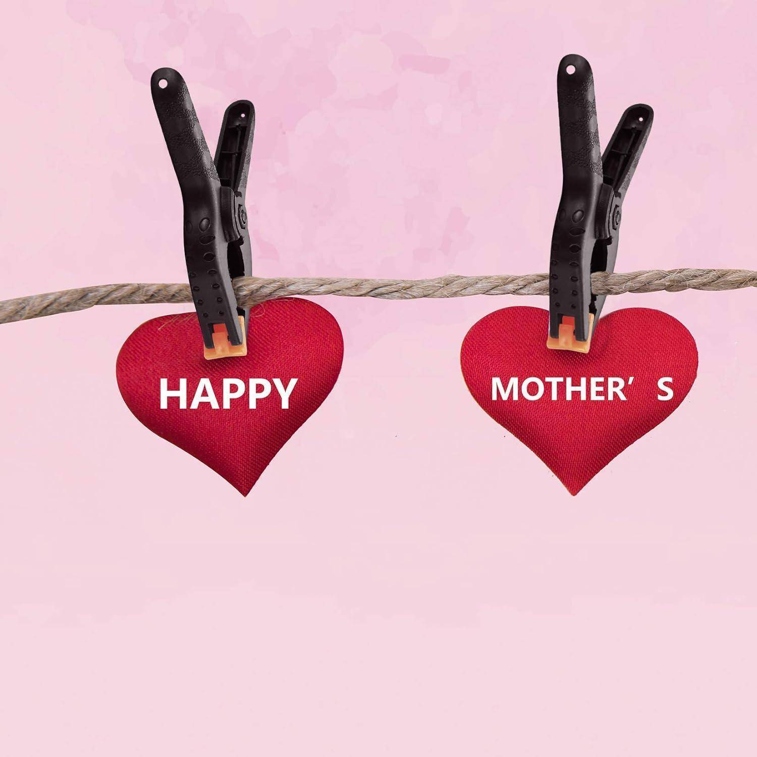 Zwei rote Herzen an einem Seil, jeweils mit einer schwarzen Wäscheklammer befestigt. Das linke Herz trägt die Aufschrift ‚HAPPY', und das rechte Herz trägt die Aufschrift ‚MOTHER'S'.