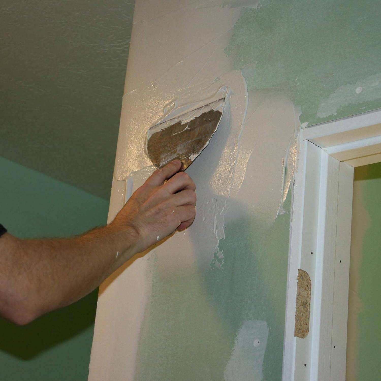 Eine Person repariert eine Wand, indem sie mit einer Spachtel Putz aufträgt und damit einen beschädigten Bereich in der Nähe des Türrahmens abdeckt.