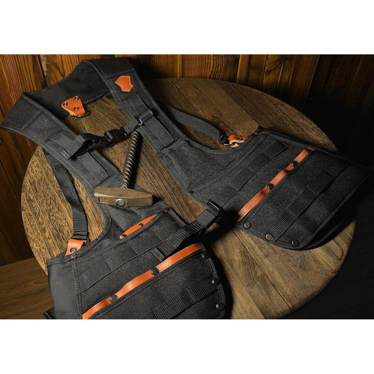Schwarze taktische Weste mit orangefarbenen Akzenten, die auf einem runden Holztisch platziert ist, begleitet von einem Hammer.