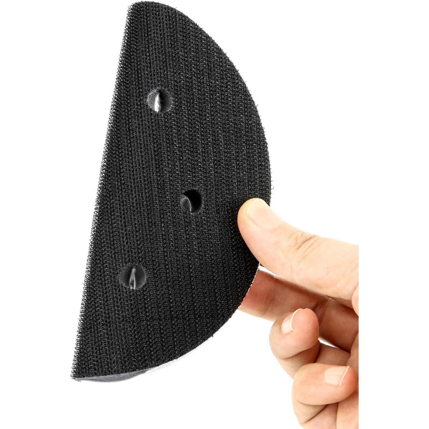 Eine Hand, die einen schwarzen, halbkreisförmigen Klettverschluss-Pad mit drei Löchern hält, der zum Befestigen an Oberflächen verwendet wird, vor einem weißen Hintergrund.