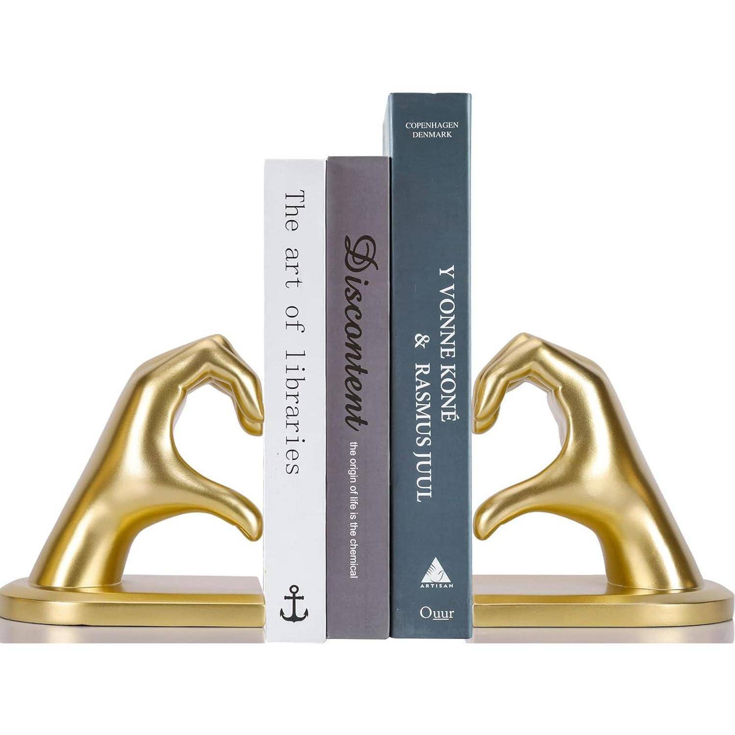 Zwei goldene, handförmige Buchstützen, die drei Bücher halten: ‚Die Kunst der Bibliotheken', ‚Unzufriedenheit' und ‚Yvonne Koné & Rasmus Stuhl'.