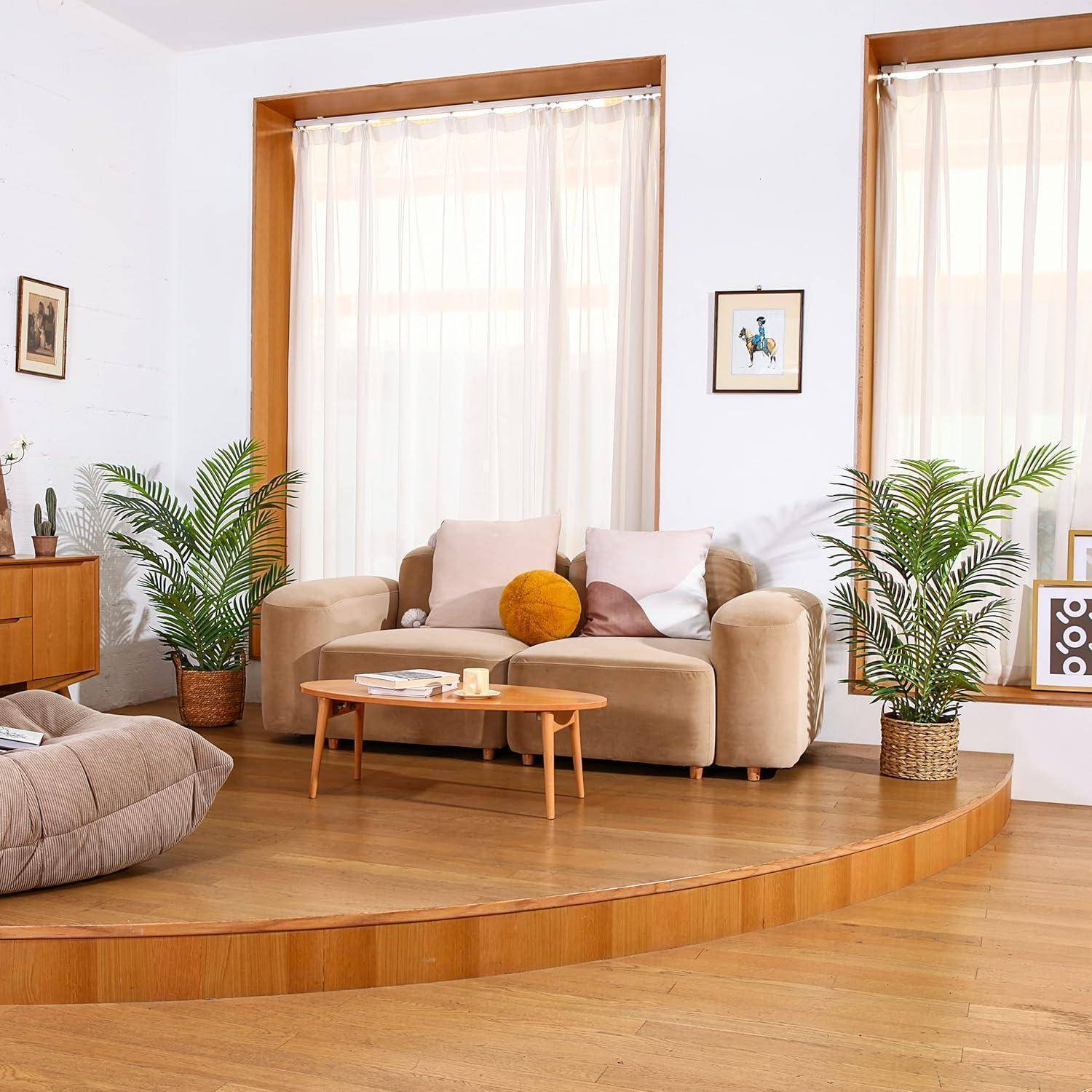 Ein gemütliches Wohnzimmer zeigt ein beiges Sofa mit Kissen, einen runden Holzkaffeetisch, Zimmerpflanzen und ein großes Fenster mit transparenten Gardinen.
