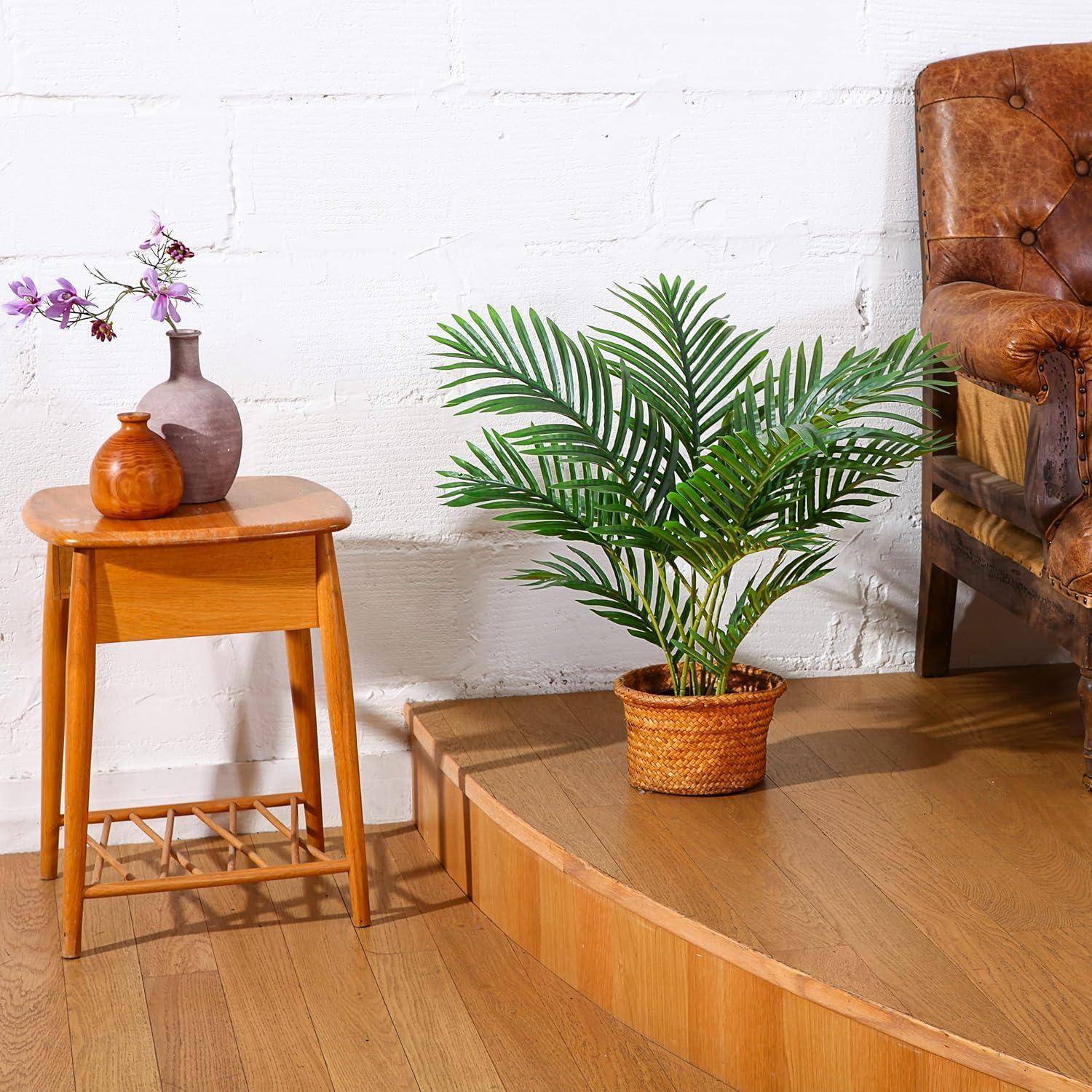Ein kleiner Holztisch mit einer Vase mit violetten Blumen und einer orangefarbenen Vase steht neben einem Flechtkorb mit einer Topfpalme auf einer Holzplattform.