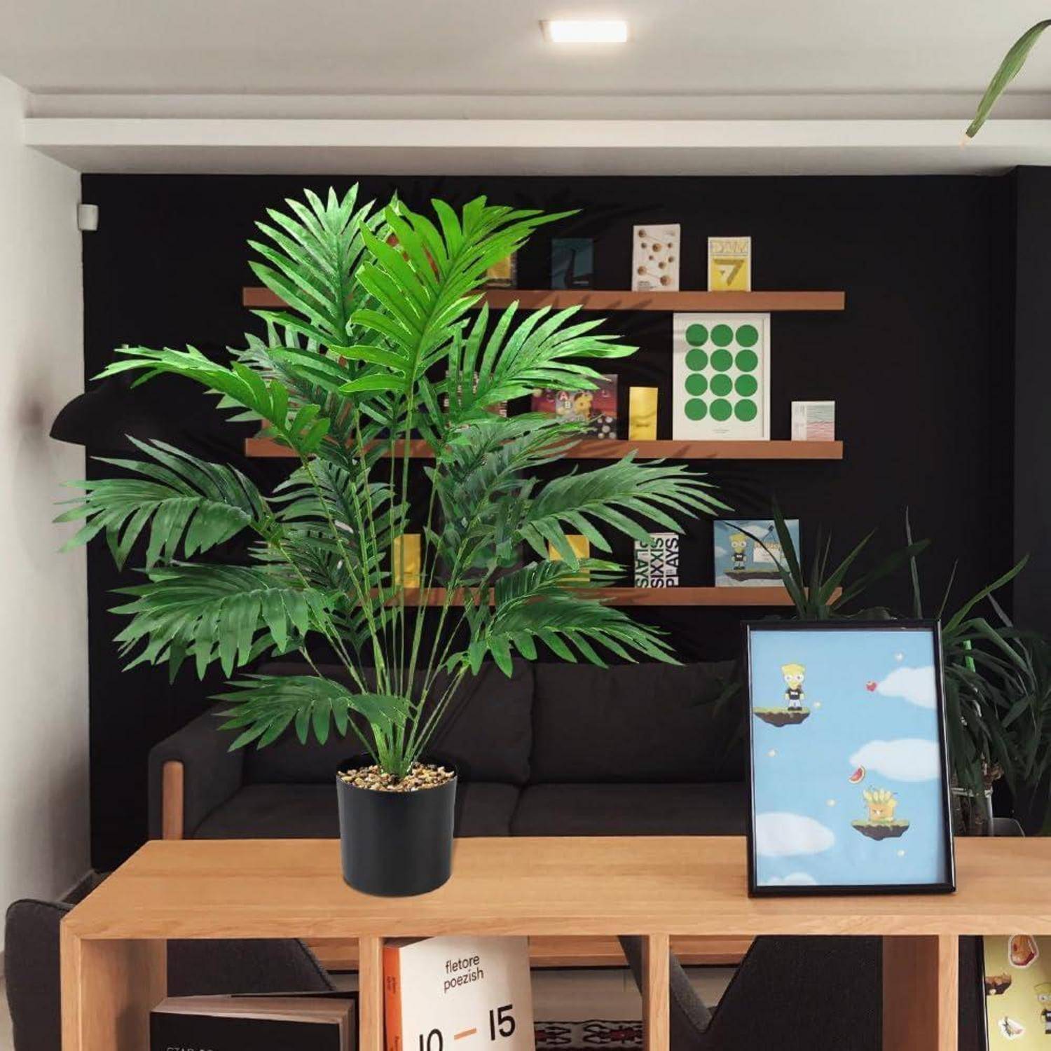 Ein moderner Wohnbereich zeigt ein schwarzes Sofa mit verschiedenen Zierbüchern und Kunstwerken auf Regalen. Eine große Topfpflanze steht auf einem Holztisch.