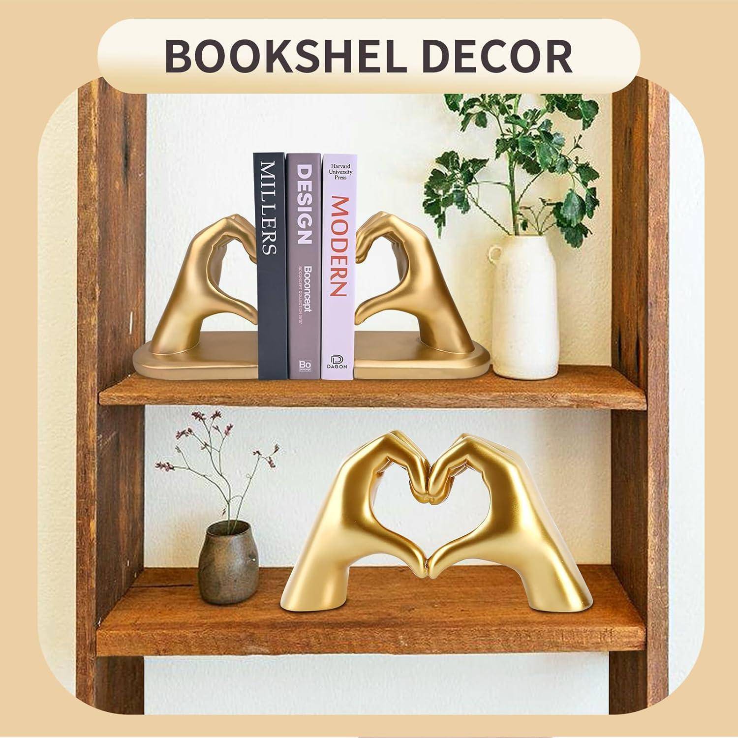 Ein hölzernes Bücherregal zeigt Bücher zwischen zwei goldenen, handförmigen Buchstützen, die ein Herz bilden. Eine Vase mit Grünpflanzen verleiht eine dekorative Note.