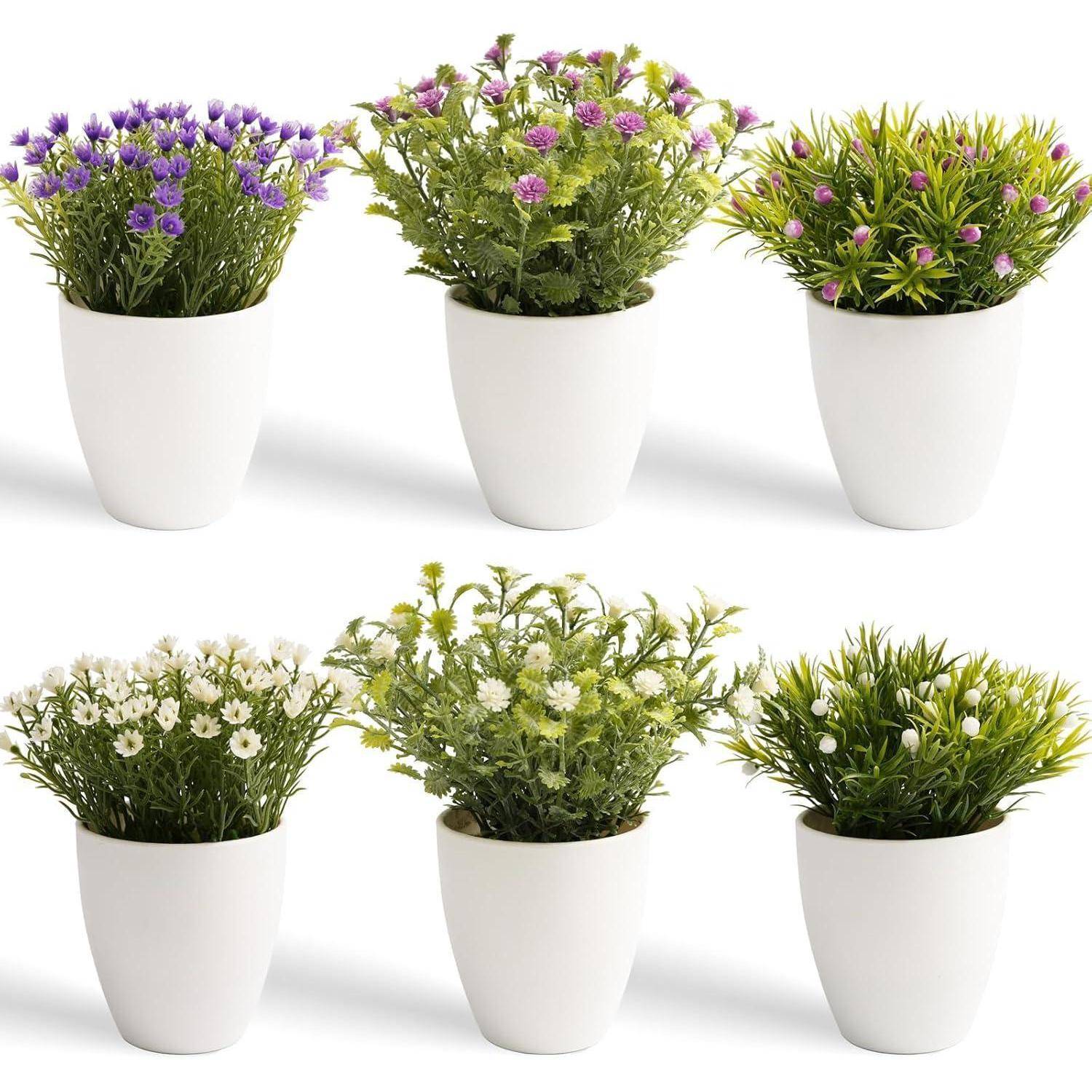 Sechs weiße Töpfe enthalten jeweils eine andere Art von künstlichen Blumen, darunter lila, rosa und weiße Blumen, angeordnet in zwei Reihen.