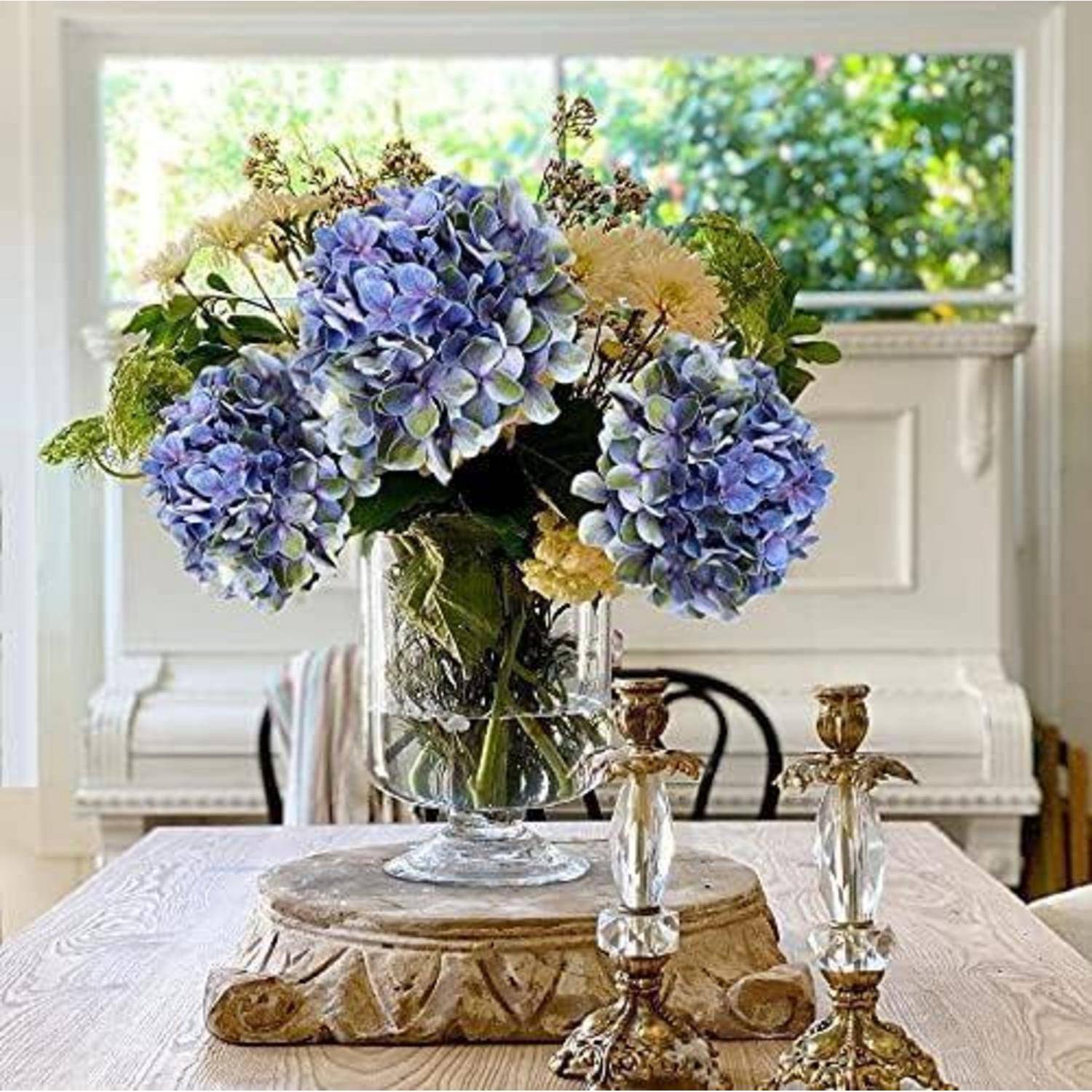 Eine klare Vase mit blauen und weißen Hortensien steht auf einem Holztisch, umgeben von zwei dekorativen Kerzenhaltern, in einem hellen Raum.