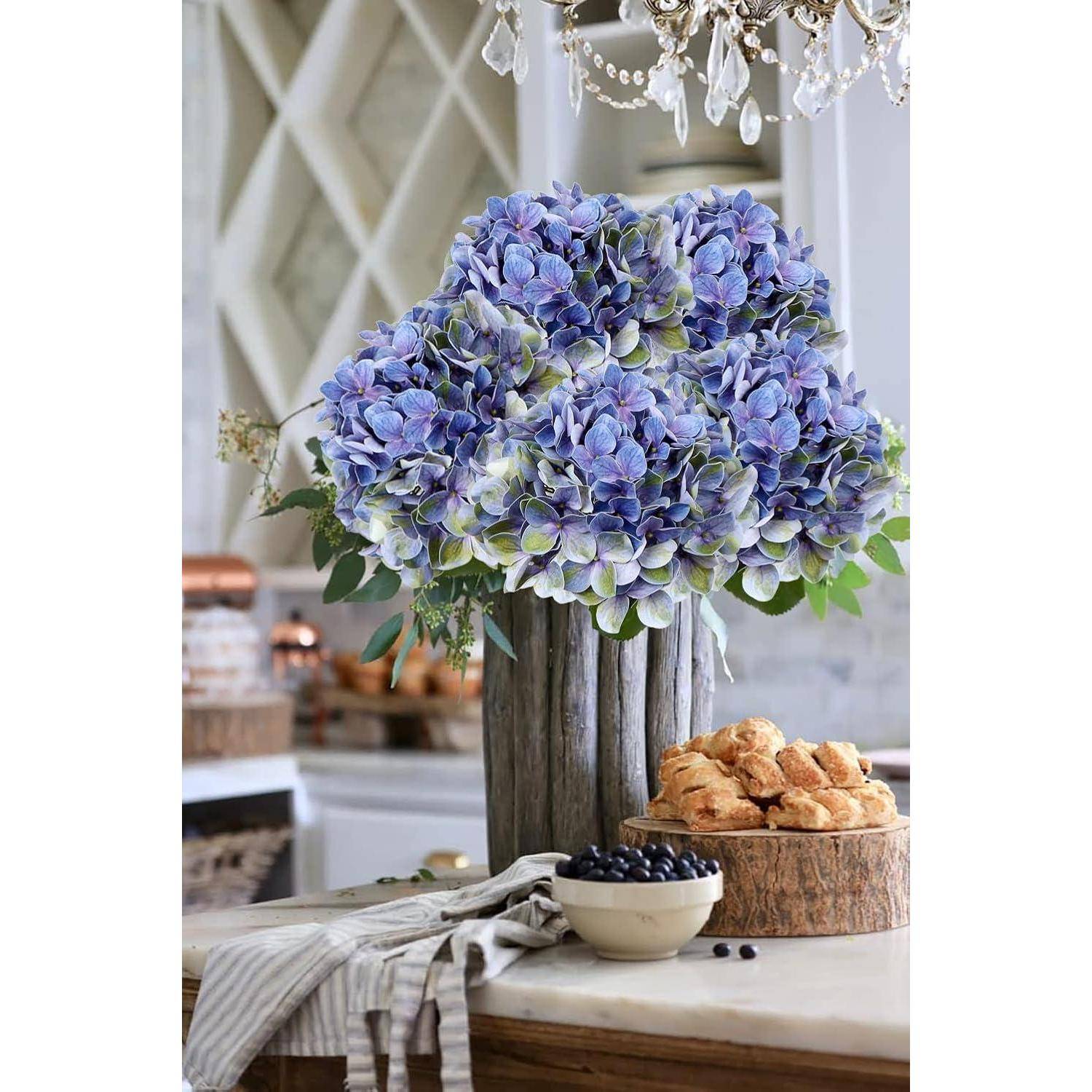 Eine Vase mit blauen Hortensien auf einem Küchentisch mit einer Schüssel voller Blaubeeren und Croissants, unter einem dekorativen Kronleuchter.