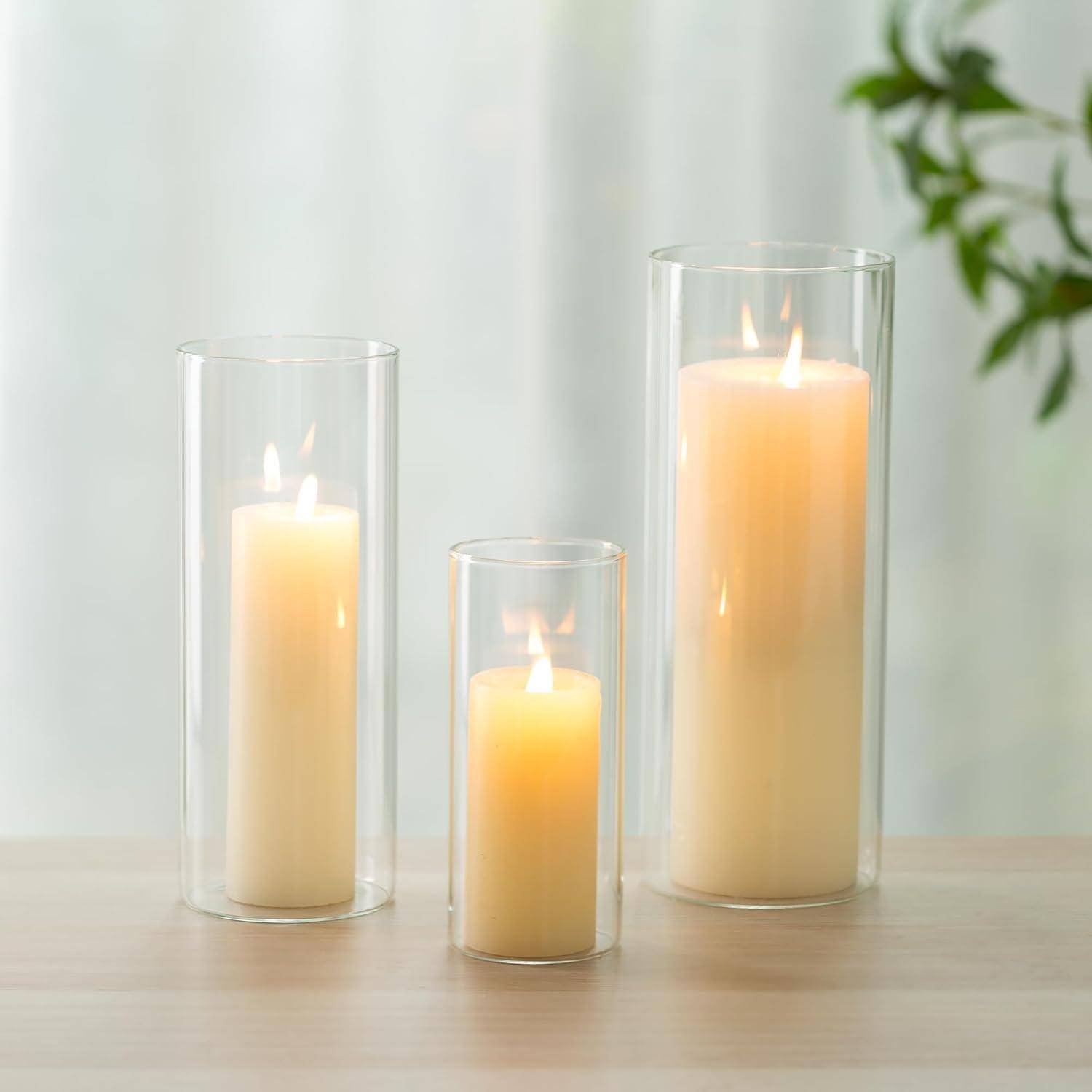 Drei beleuchtete Kerzen in Säulenform in transparenten Glashaltern unterschiedlicher Höhe stehen auf einer Holzoberfläche. Im Hintergrund ist teilweise eine grüne Pflanze zu sehen.