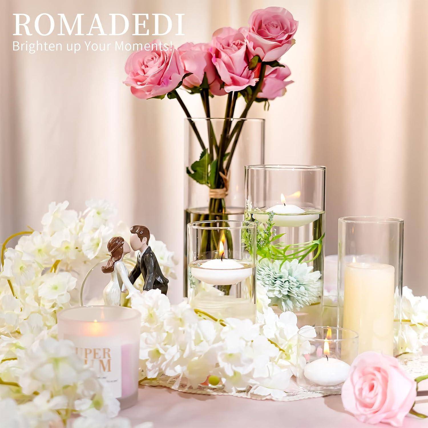Eine Hochzeitsszenerie mit rosa Rosen in einer Vase, schwimmenden Kerzen und einer Braut- und Bräutigam-Figur, vor einem weißen Blumenhintergrund.