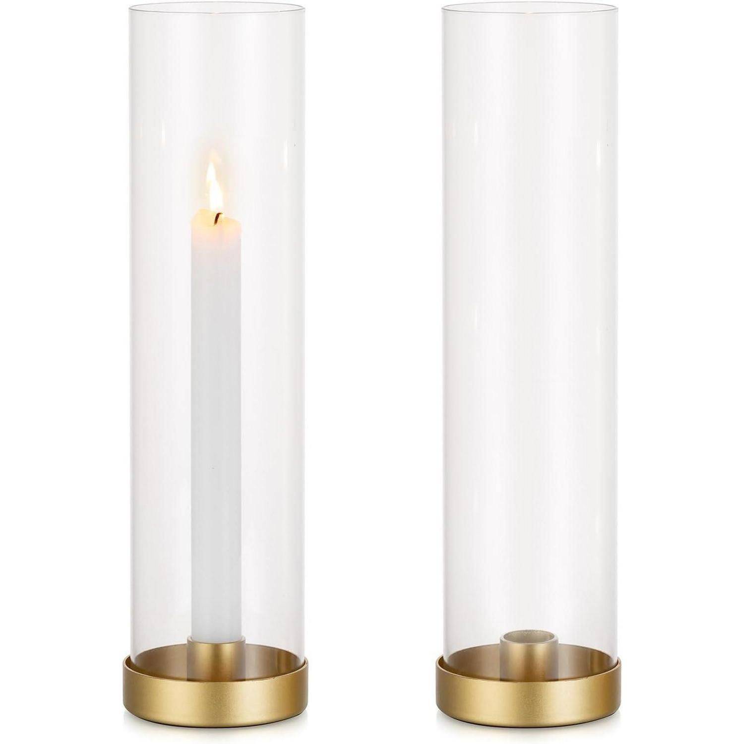 Zwei hohe Glaswindlichter mit goldenen Füßen; eines enthält eine brennende weiße Stabkerze, das andere ist leer.
