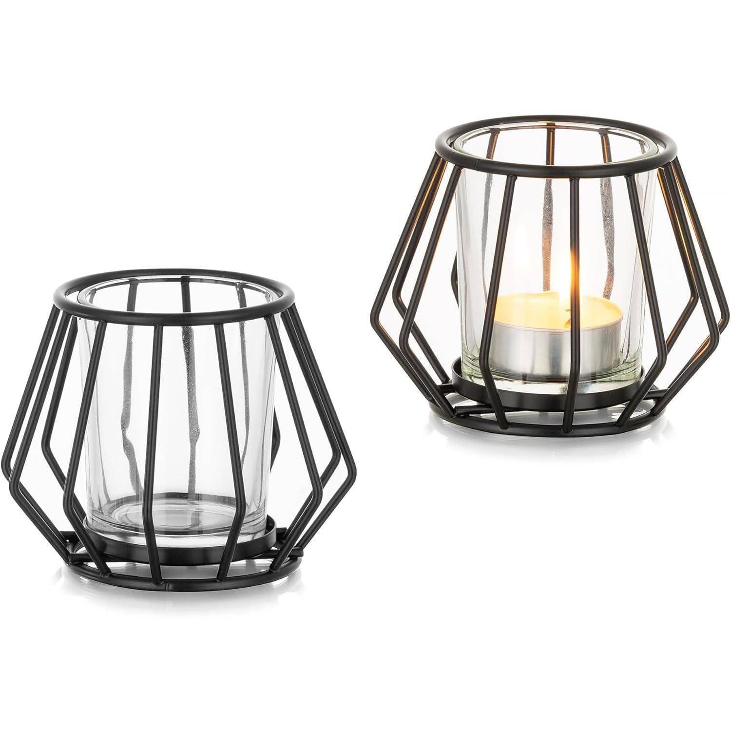 Zwei Kerzenhalter aus Metall, einer davon mit einer brennenden Kerze im Inneren, die ein warmes Glühen erzeugt. Die Halter weisen ein geometrisches, käfigartiges Design auf.