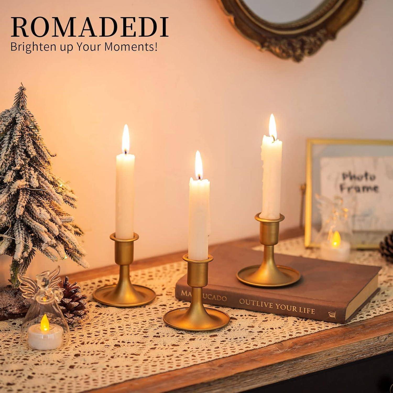 Drei beleuchtete Kerzen in goldenen Haltern auf einem mit Deckchen bedeckten Tisch in der Nähe eines kleinen künstlichen Baumes und einiger Bücher. Wandschild zeigt: 'ROMADEDI Erhelle deine Momente!