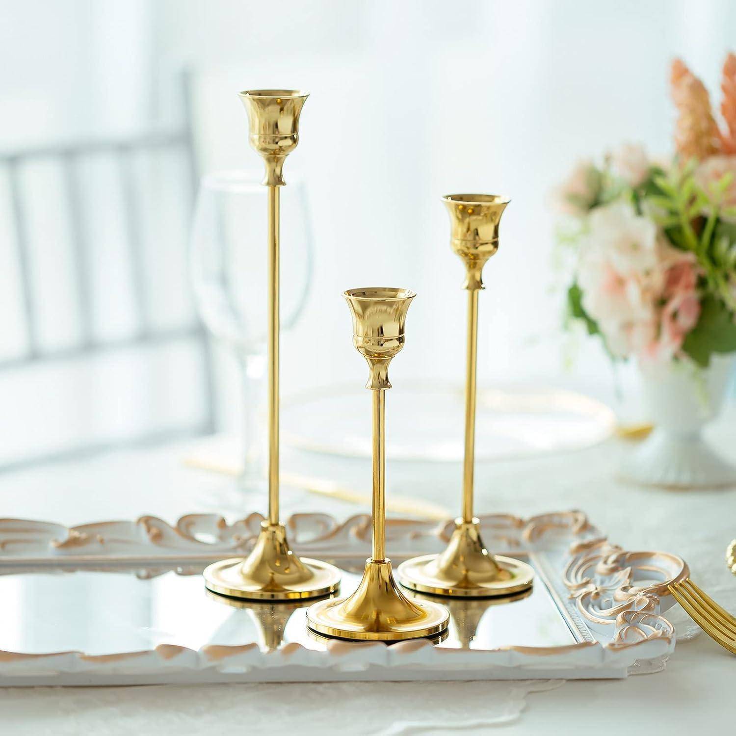 Goldene Kerzenleuchter auf einem Spiegeltablett mit unscharfen Blumen im Hintergrund, die eine elegante Tischdekoratmosphäre schaffen.