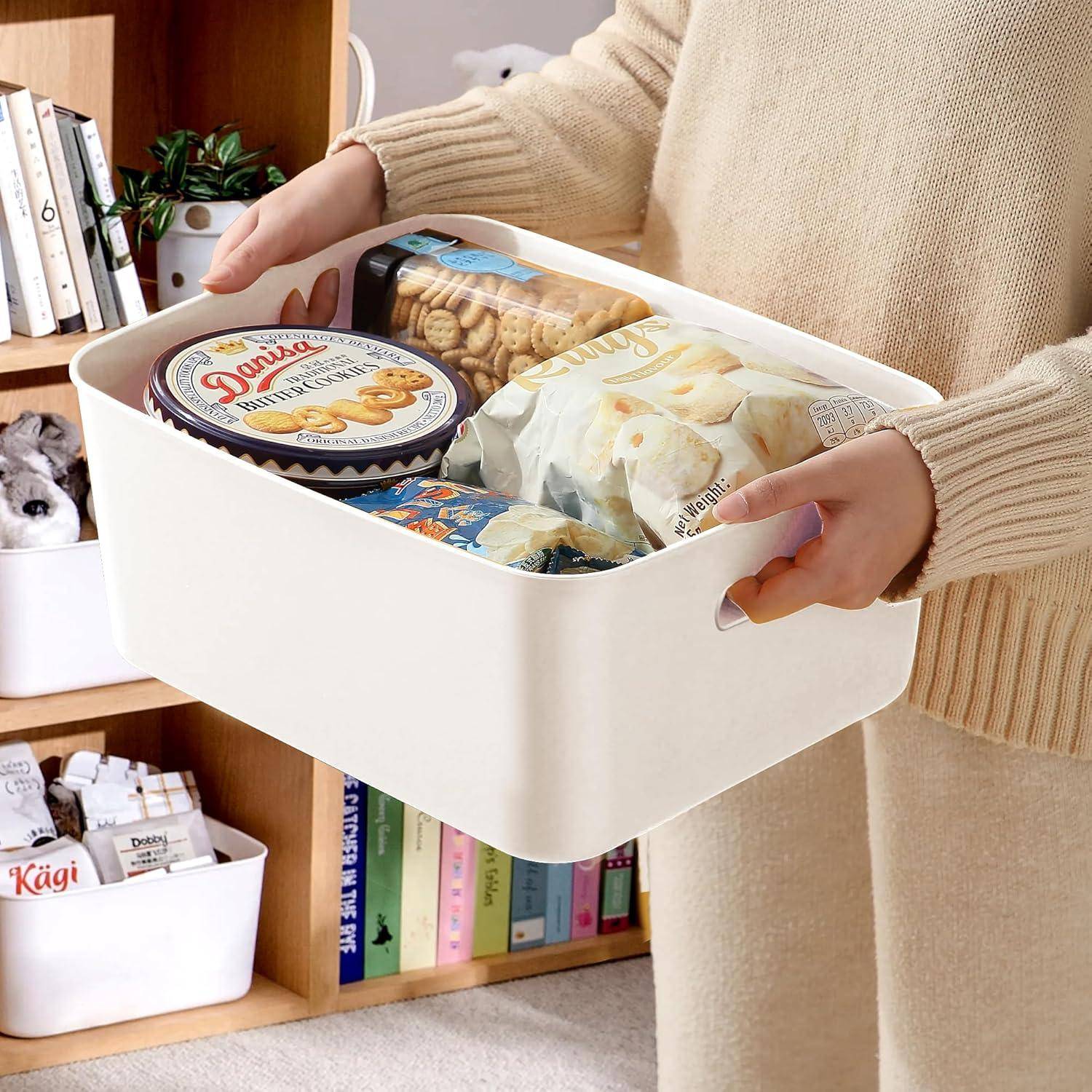 Eine Person hält eine weiße Aufbewahrungsbox, die mit Snacks und verpackten Lebensmitteln gefüllt ist. Bücher und Aufbewahrungsbehälter sind im Hintergrund sichtbar.