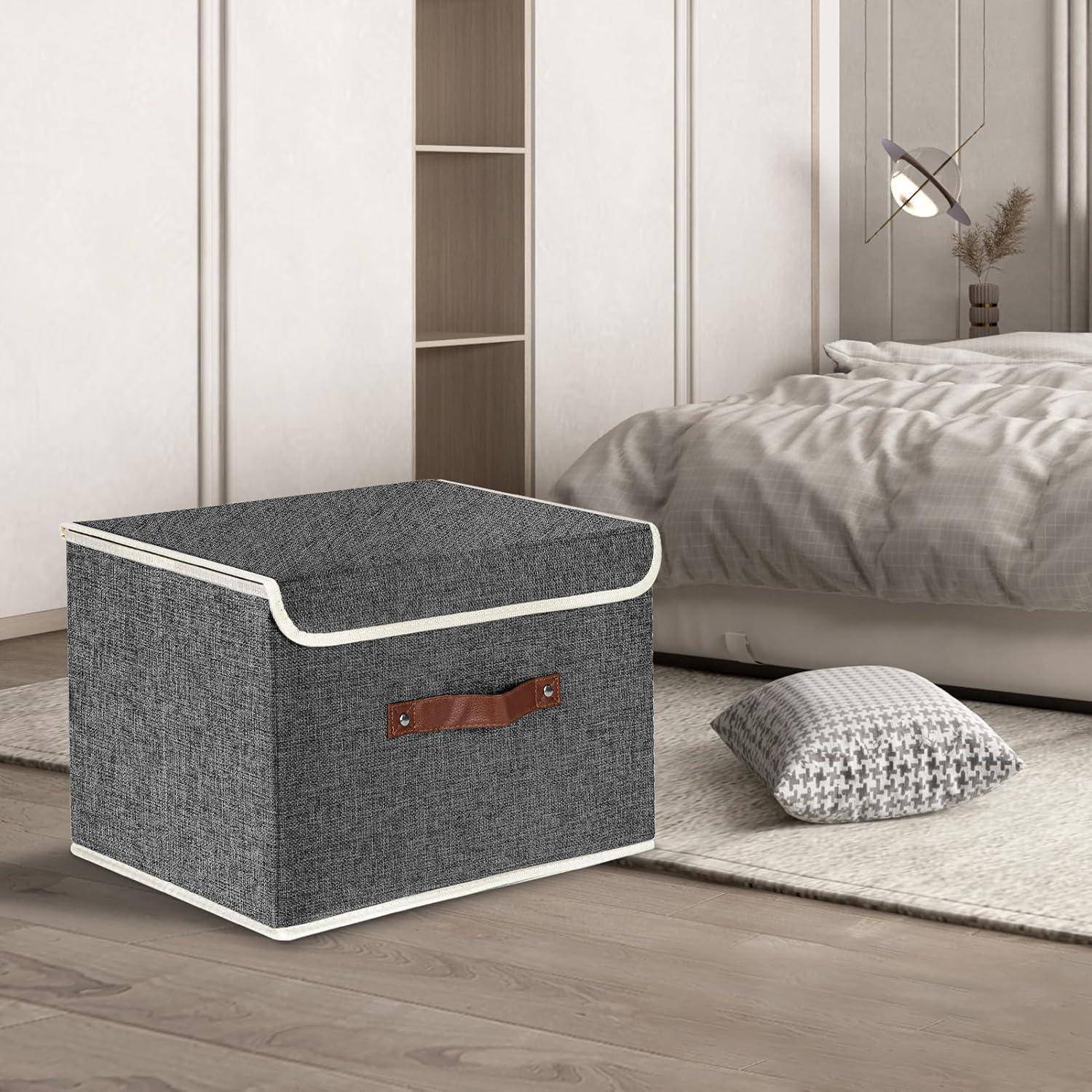 Eine graue Aufbewahrungsbox aus Stoff mit einem braunen Griff steht auf einem Holzboden neben einem Bett mit grauen Laken und einem gewebten Kissen.