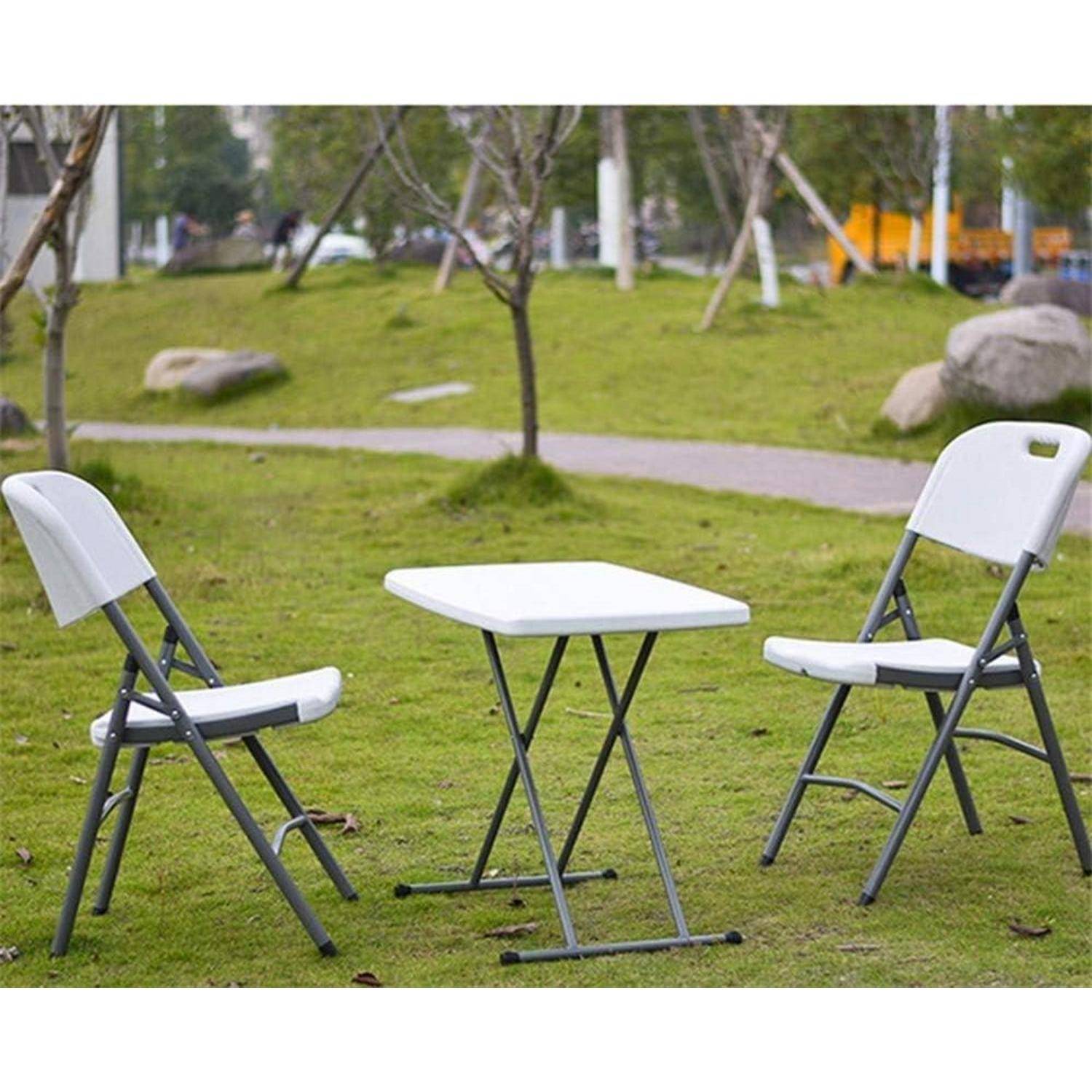 Außenszene mit zwei weißen Klappstühlen und einem kleinen quadratischen Tisch auf einer Grasfläche. Im Hintergrund ist ein Weg und sind Bäume zu sehen.