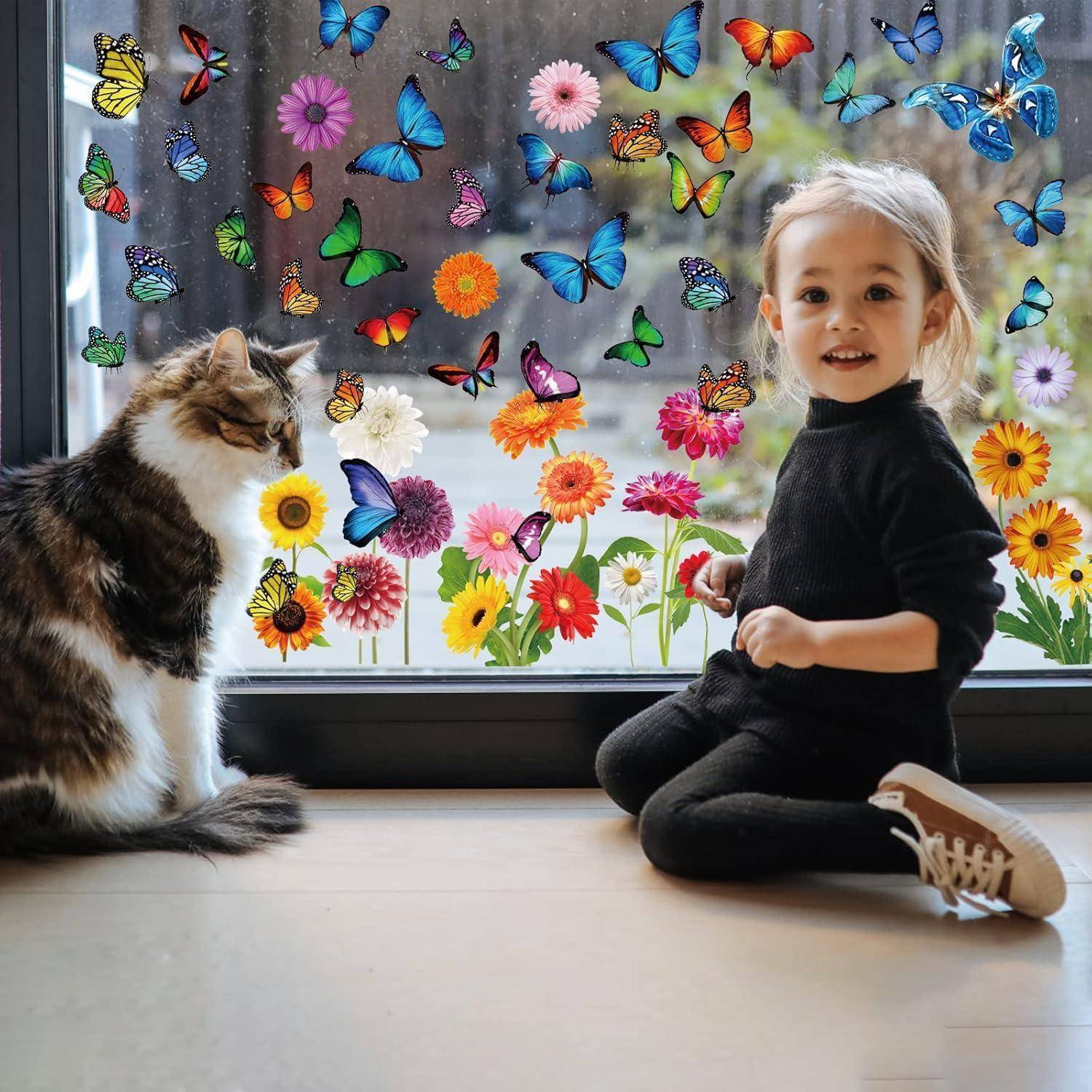 Ein junges Kind in Schwarz sitzt an einem Fenster, das mit bunten Schmetterlingen und Blumen geschmückt ist, und lächelt eine getigerte Katze an, die neben ihm sitzt.