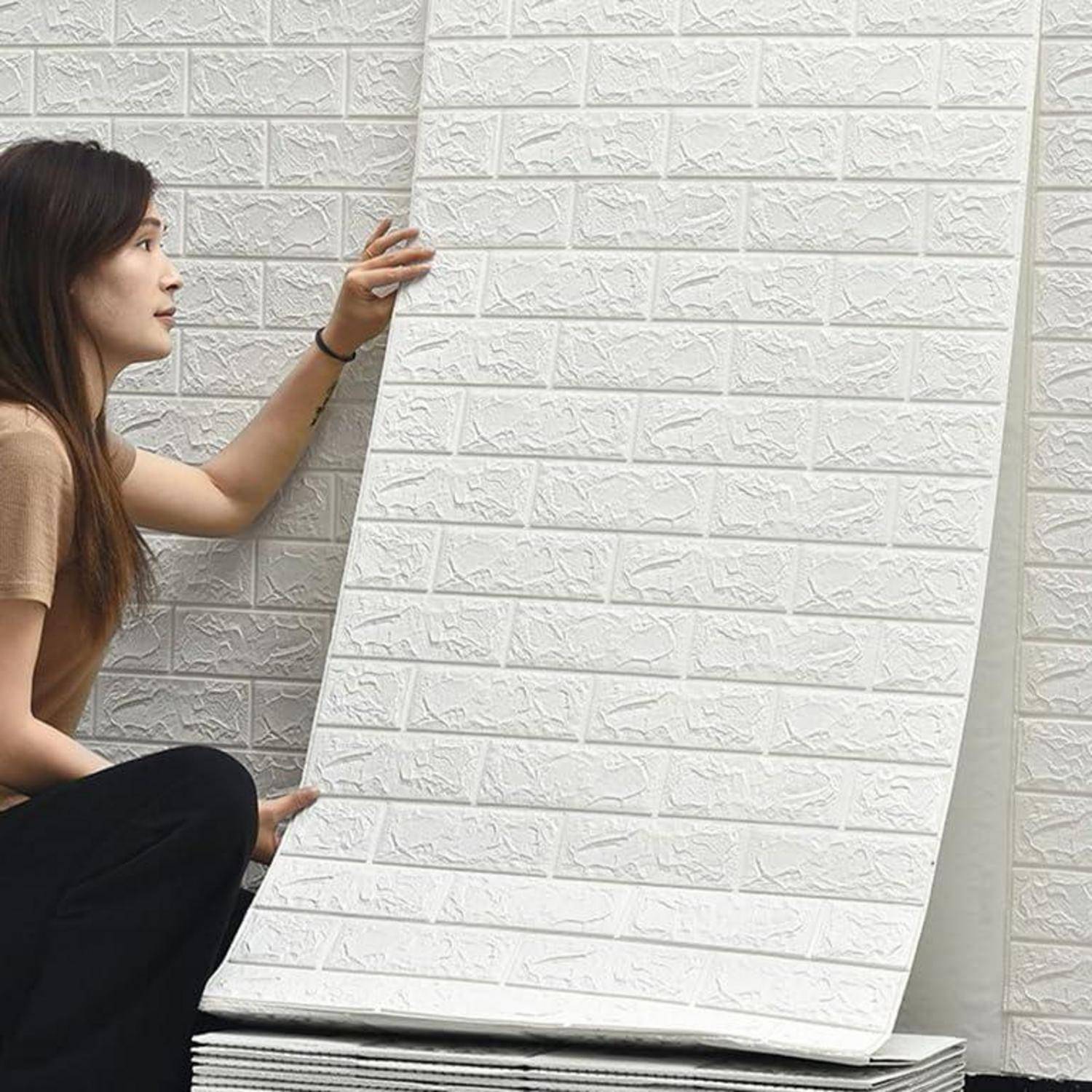 Eine Frau sitzt neben einem Stapel weißer 3D-Wandpaneele mit Ziegelsteinmuster, wodurch deren Textur und Design präsentiert werden.