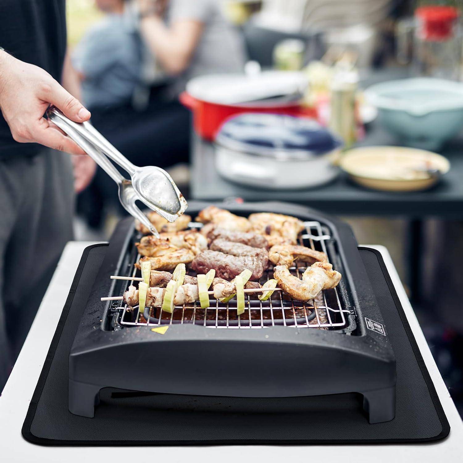 Eine Person grillt Fleisch- und Gemüsespieße auf einem elektrischen Tischgrill drinnen, wobei im unscharfen Hintergrund ein Tisch mit Geschirr und Küchenutensilien zu sehen ist.