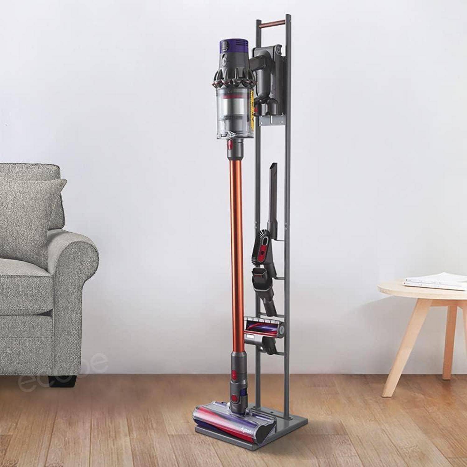 Ein kabelloser Staubsauger auf einem Ständer neben einem grauen Sofa und einem Holzseitentisch vor einer hellgefärbten Wand.