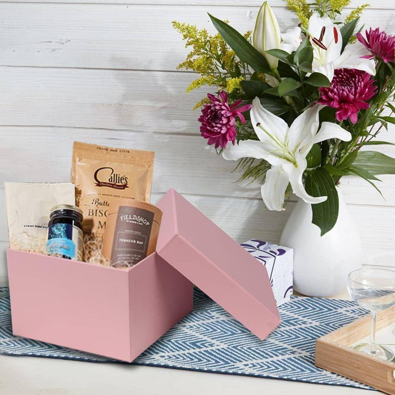 Rosa Geschenkbox mit Delikatessen, darunter Kekse und Kaffee, neben einer Vase mit verschiedenen Blumen auf einem Tisch mit blauem Läufer.