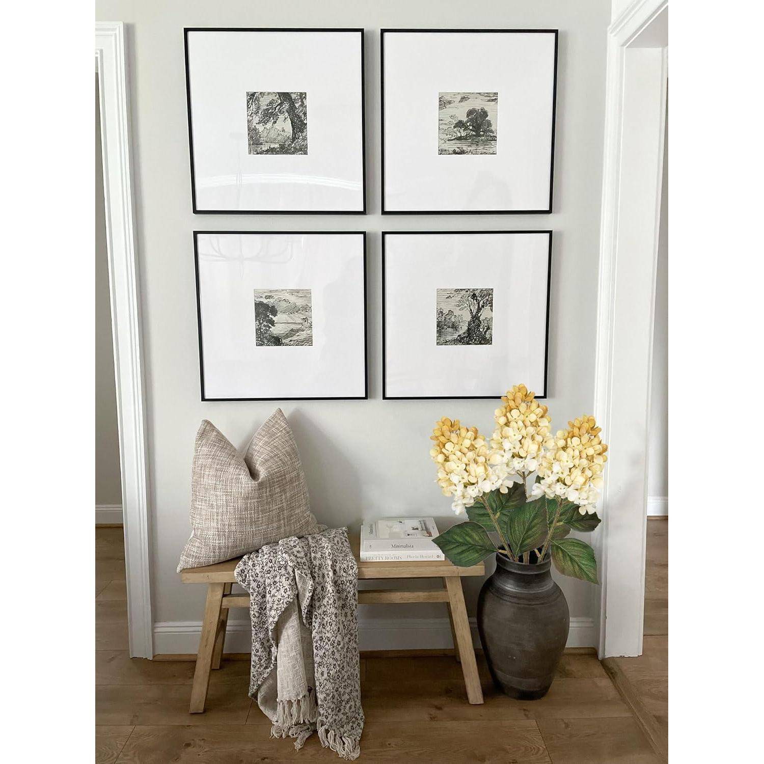 Eine Holzbank unter vier gerahmten Kunstwerken an einer weißen Wand. Ein dekoratives Kissen und eine Decke liegen auf der Bank, neben einer Vase mit Blumen.