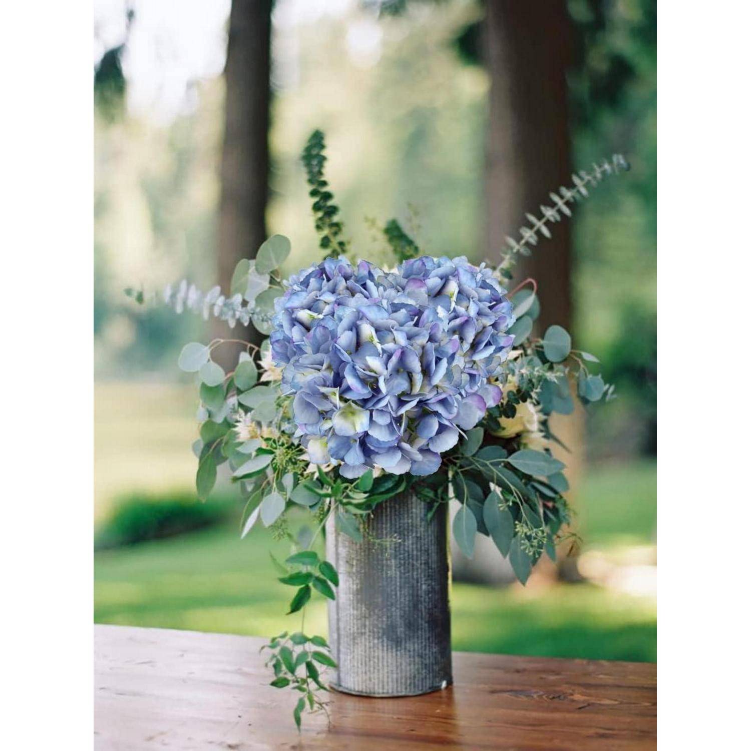 Ein Strauß blauer Hortensien und Blattwerk in einer Metalvase steht auf einem Holztisch im Freien mit unscharfen Bäumen im Hintergrund.