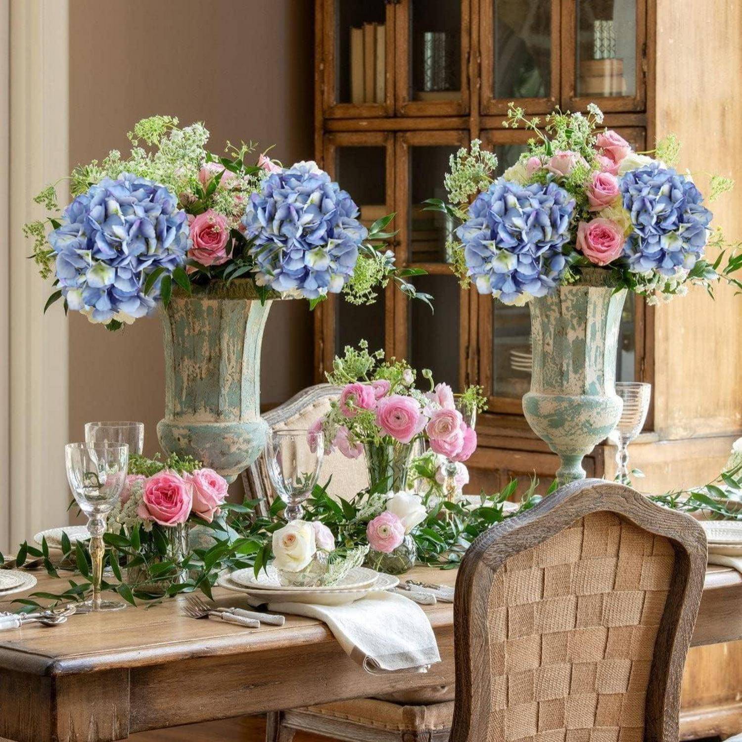 Ein Esstisch elegant gedeckt mit hohen Vasen aus blauen und rosa Hortensien, umgeben von Grünpflanzen, die eine frische, einladende Atmosphäre schaffen.