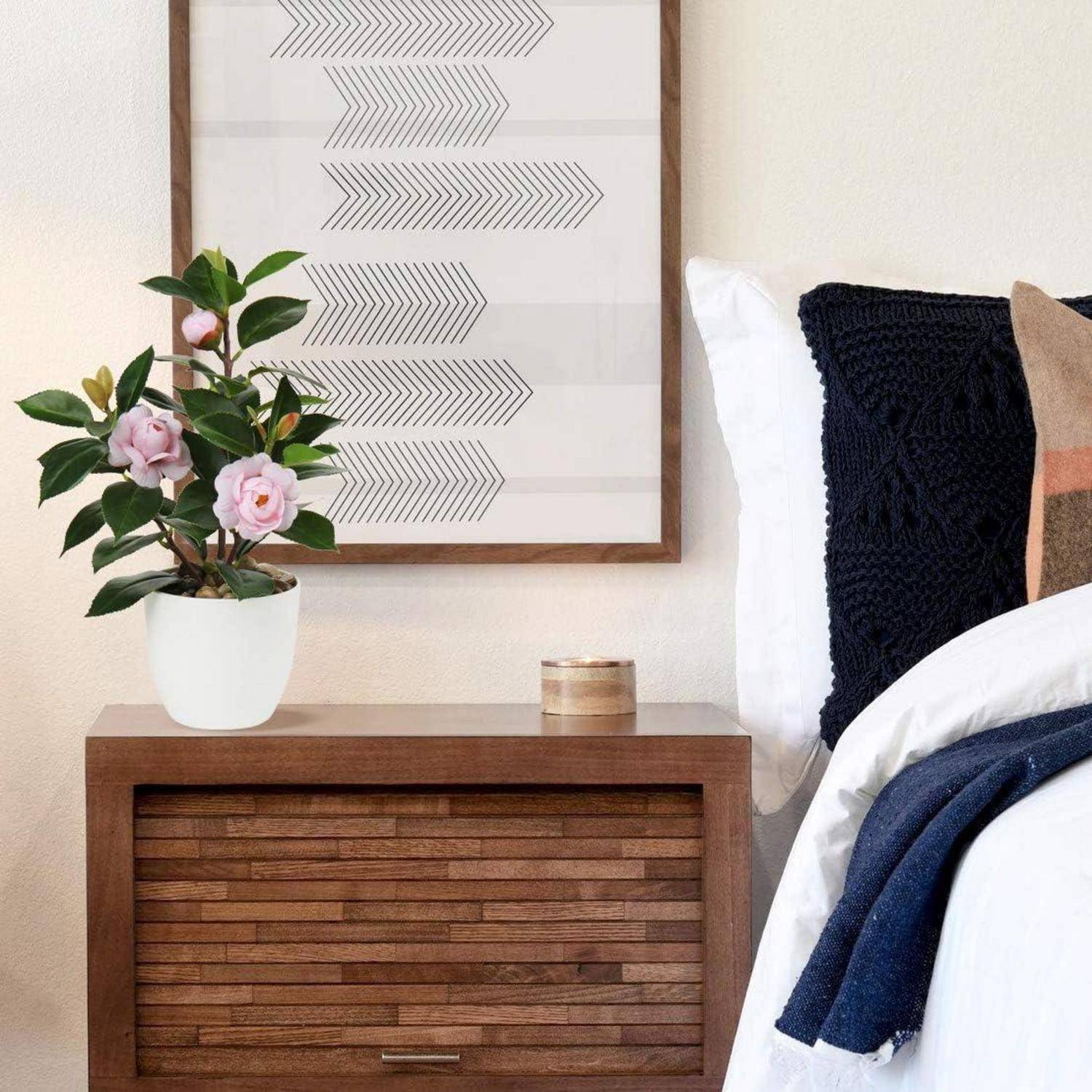 Ein modernes Schlafzimmer mit einem Holznachttisch, einer Topfpflanze mit rosa Blüten, einem geometrischen Wandkunstwerk und einem Teil eines ordentlich gemachten Bettes.