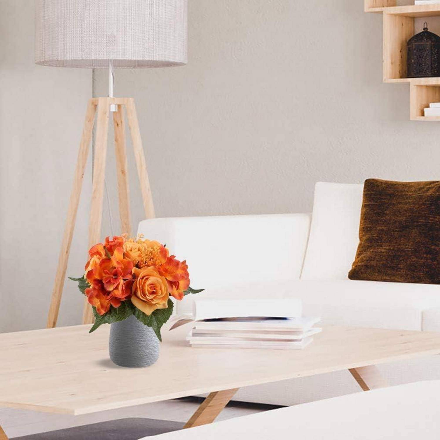Ein Wohnzimmer mit einem weißen Sofa und braunem Kissen, einem hellholzfarbenen Couchtisch und einer Dreibeinlampe. Eine Vase mit leuchtend orangefarbenen Blumen steht auf dem Tisch.