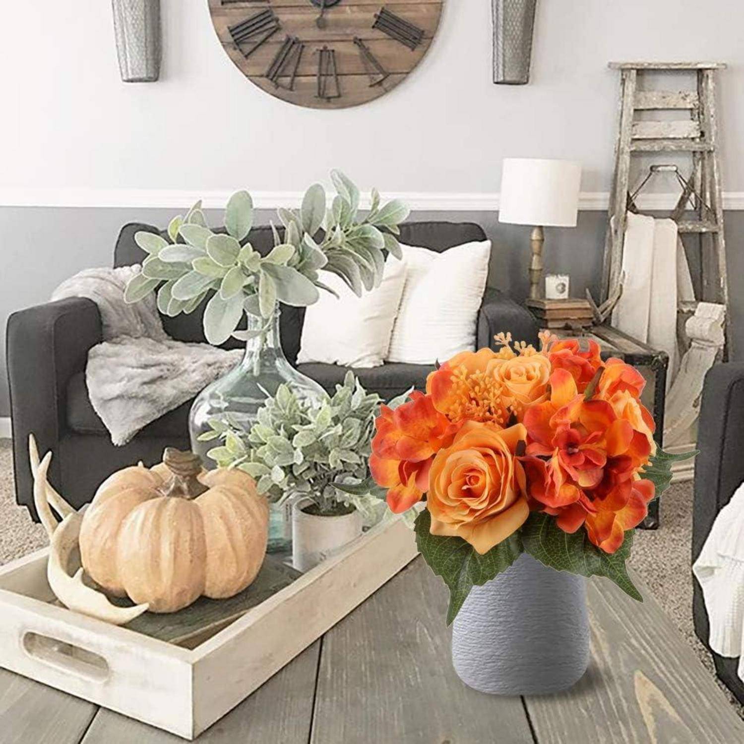 Wohnzimmer mit einem grauen Sofa, Holzkaffeetisch mit Kürbissen und Blumen, Wanduhr darüber, wodurch eine gemütliche Herbststimmung entsteht.