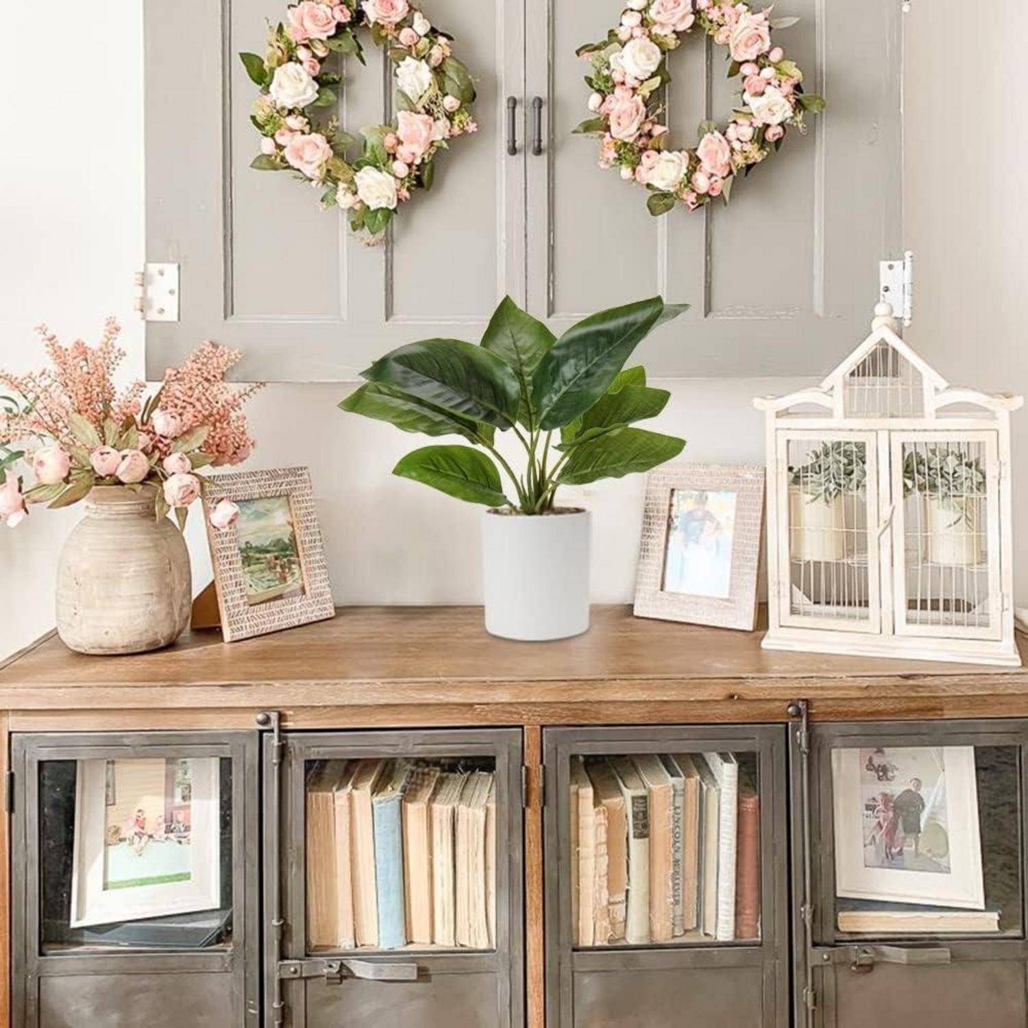 Ein rustikales Regal beherbergt alte Bücher, gerahmte Fotos, eine Topfpflanze und dekorative Blumenkränze, die darüber hängen und eine Vintage-Ästhetik erschaffen.
