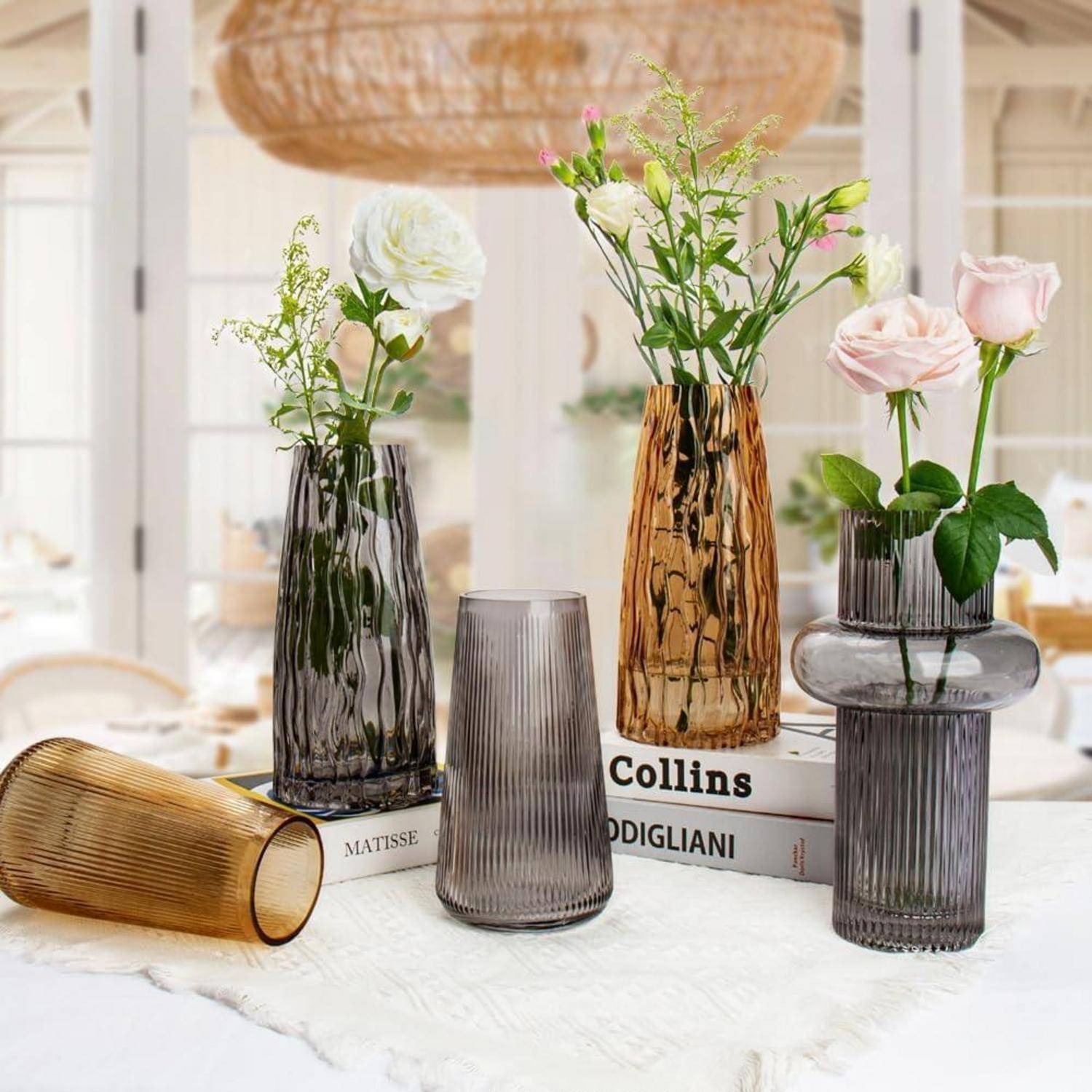 Verschiedene dekorative Glasvasen mit Blumen, die auf gestapelten Büchern platziert sind, in einem sanft beleuchteten Raum, der eine elegante und ruhige Atmosphäre schafft.