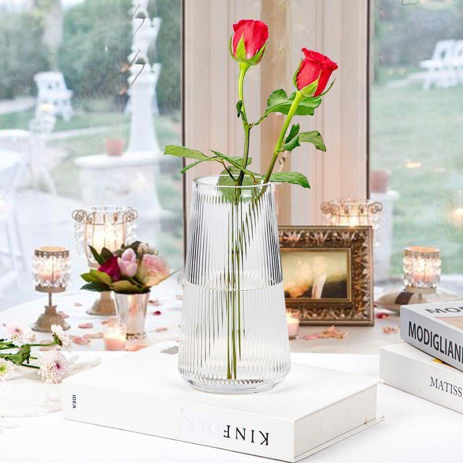 Eine weiße Vase mit zwei roten Rosen steht auf einem Tisch, der für eine elegante Outdoor-Zusammenkunft gedeckt ist und von Büchern und Kerzen umgeben ist.