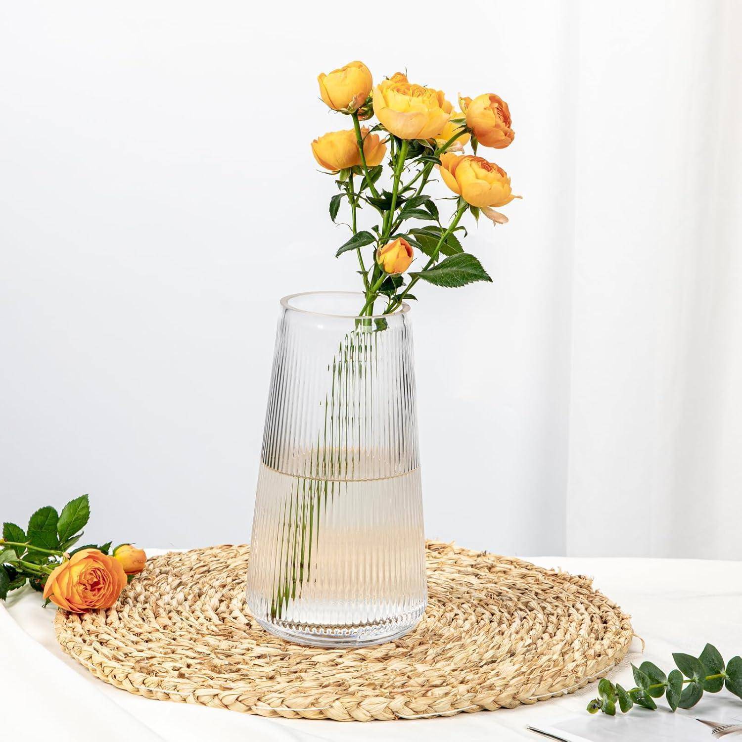 Eine klare Glasvase mit gelben Rosen auf einer geflochtenen Matte. Eine einzelne Rose liegt neben der Vase. Helle Atmosphäre.