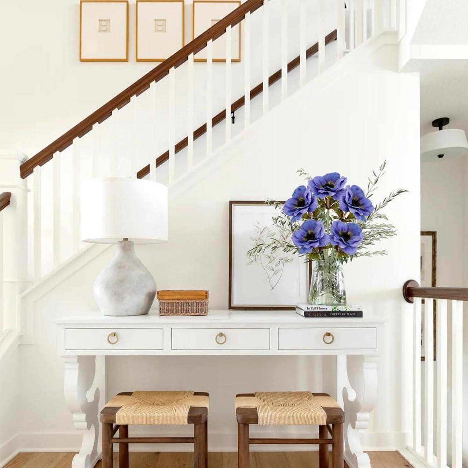 Ein heller, minimalistischer Eingangsbereich zeigt einen weißen Konsolentisch mit einer Lampe, Büchern und einer Vase mit großen blauen Blumen. Die Treppe führt dahinter nach oben.