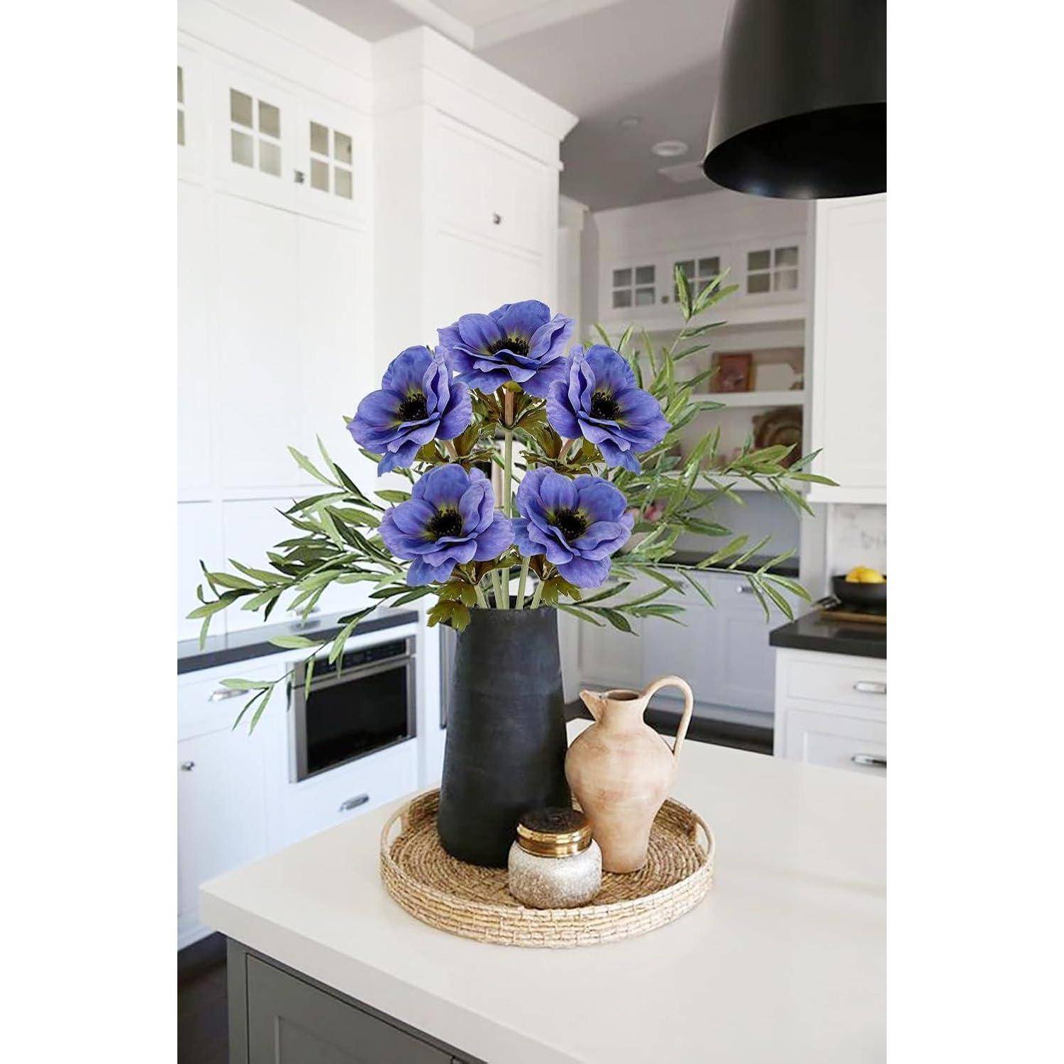 Eine schwarze Vase mit lila Blumen und grünen Blättern steht auf einer Küchenarbeitsplatte. In der Nähe befinden sich ein kleines Glas und ein Tonkrug auf einem geflochtenen Tablett.