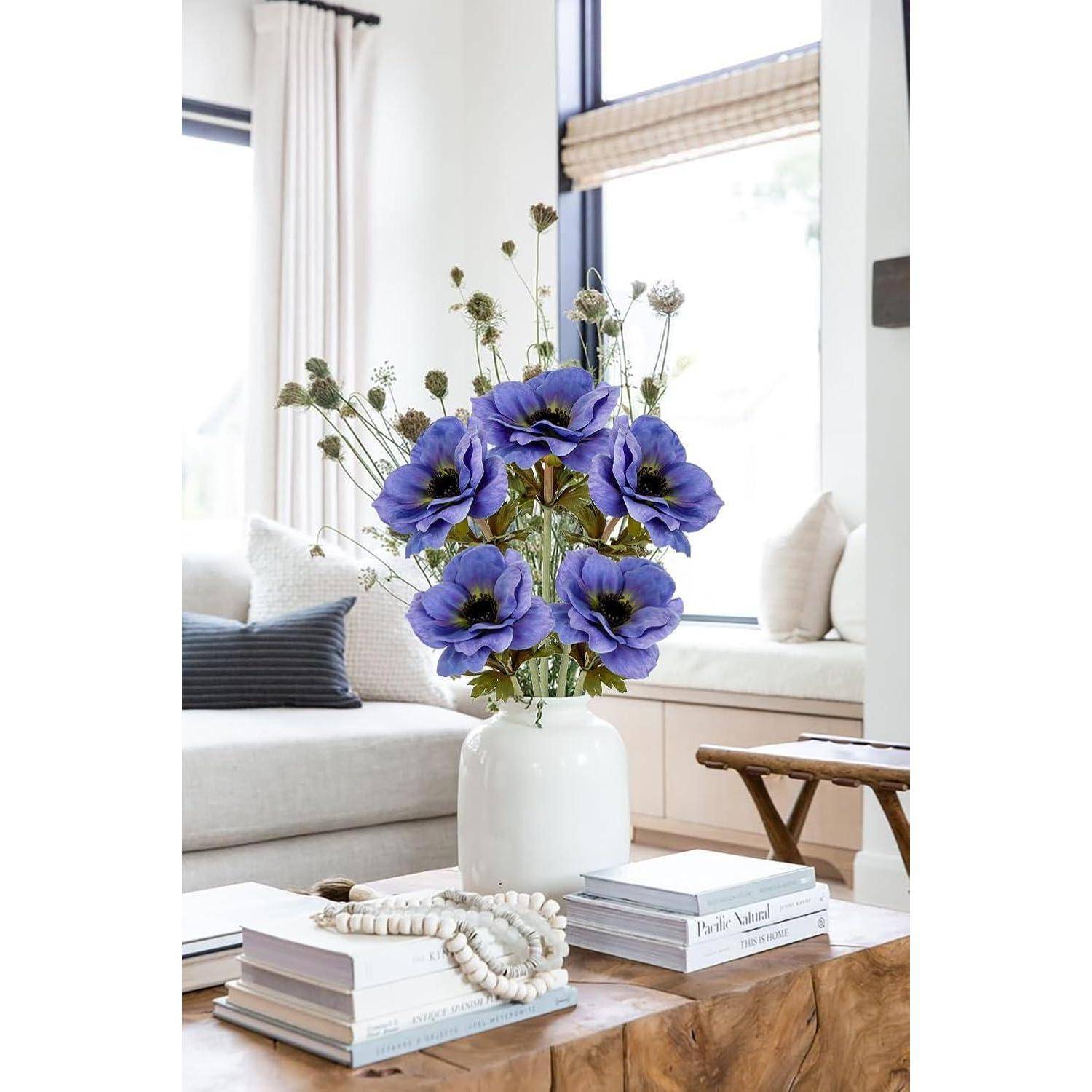 Eine Vase mit blauen Blumen steht auf einem Holztisch, umgeben von Büchern und dekorativen Perlen in einer hellen Wohnzimmeratmosphäre.
