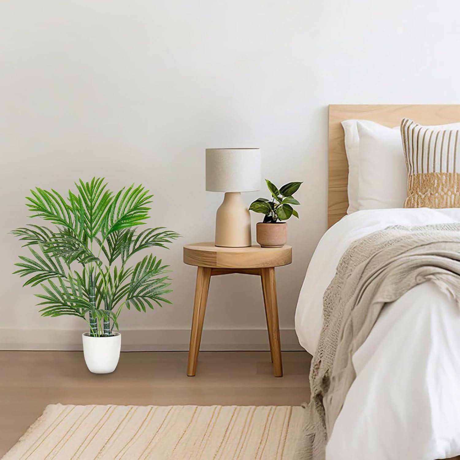Ein gemütliches Schlafzimmer mit einem weißen Bett, einem Holznachttisch mit einer Lampe, einer Topfpflanze und einem beigen Teppich auf einem Holzboden. Schlichte und ruhige Einrichtung.