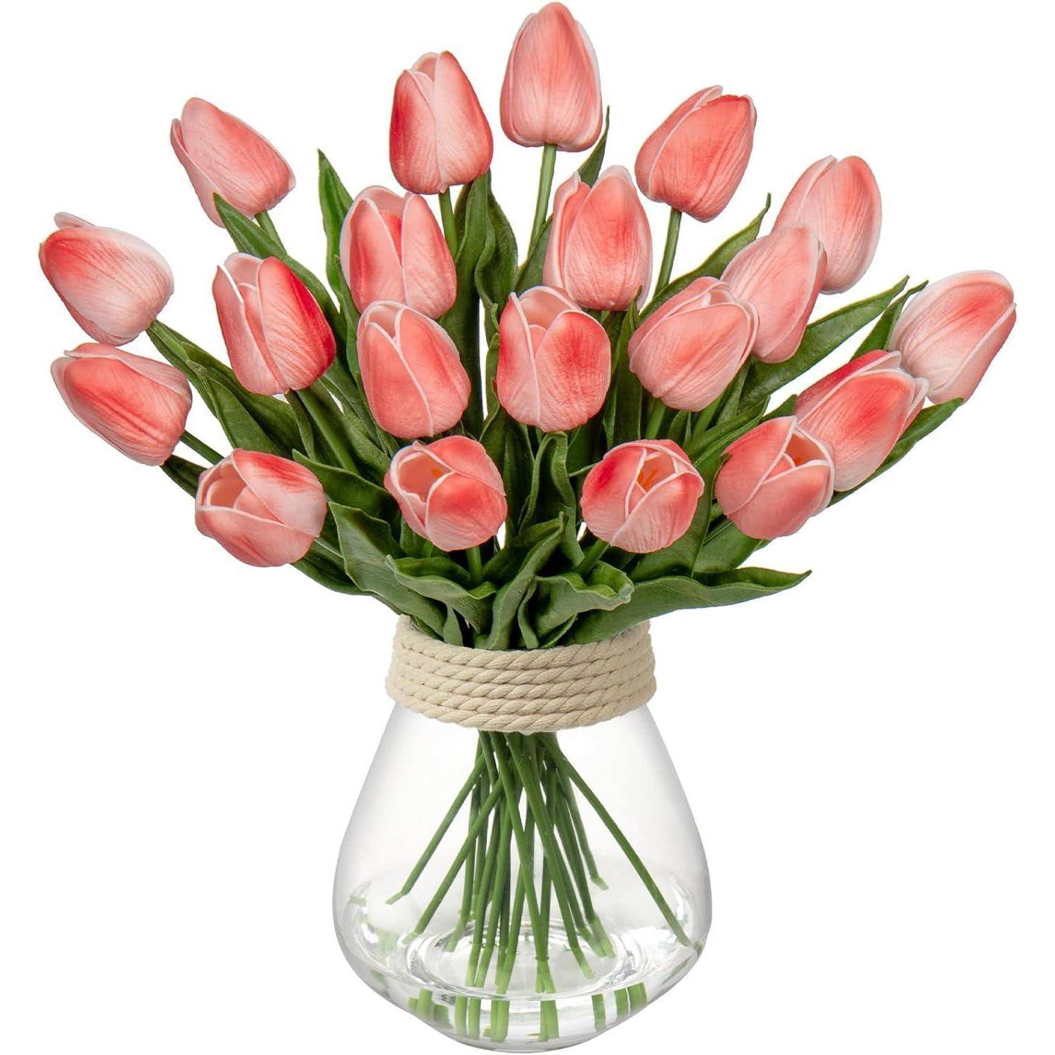 Ein Blumenstrauß aus rosa Tulpen mit grünen Blättern, in einer durchsichtigen Vase angeordnet und mit einem Seilakzent umwickelt, vor einem einfarbigen Hintergrund präsentiert.