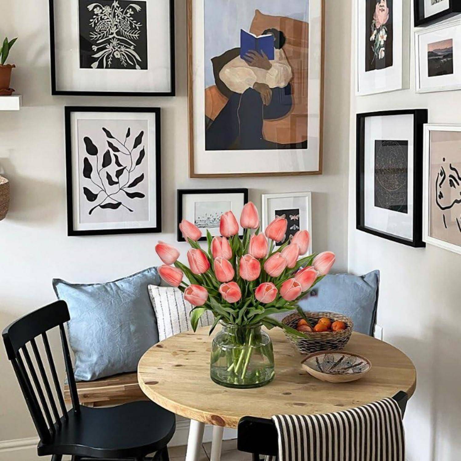 Eine gemütliche Essecke mit einem runden Holztisch, einer Vase mit rosa Tulpen und gerahmten Kunstwerken an den Wänden. Eine kleine Schale mit Orangen steht auf dem Tisch.