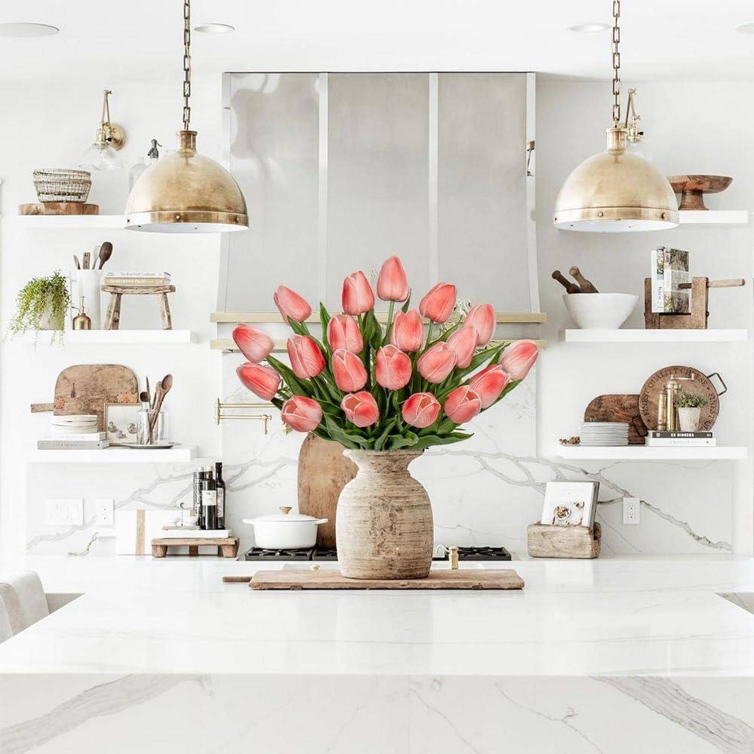 Eine moderne Küche mit weißen Marmor-Arbeitsplatten und Regalen, mit einer großen Vase mit rosa Tulpen auf der Kücheninsel, unter goldenen Pendelleuchten.