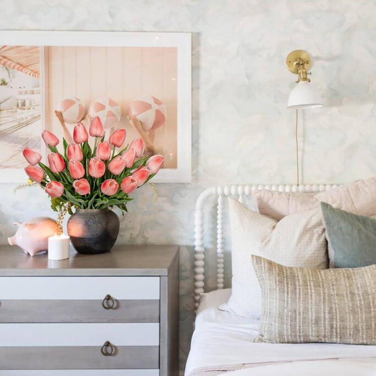 Ein Schlafzimmer mit einem helltonigen Bett, Kissen und einem Nachttisch. Eine Vase mit rosa Tulpen steht auf dem Nachttisch, und ein großes gerahmtes Foto hängt an der Wand.
