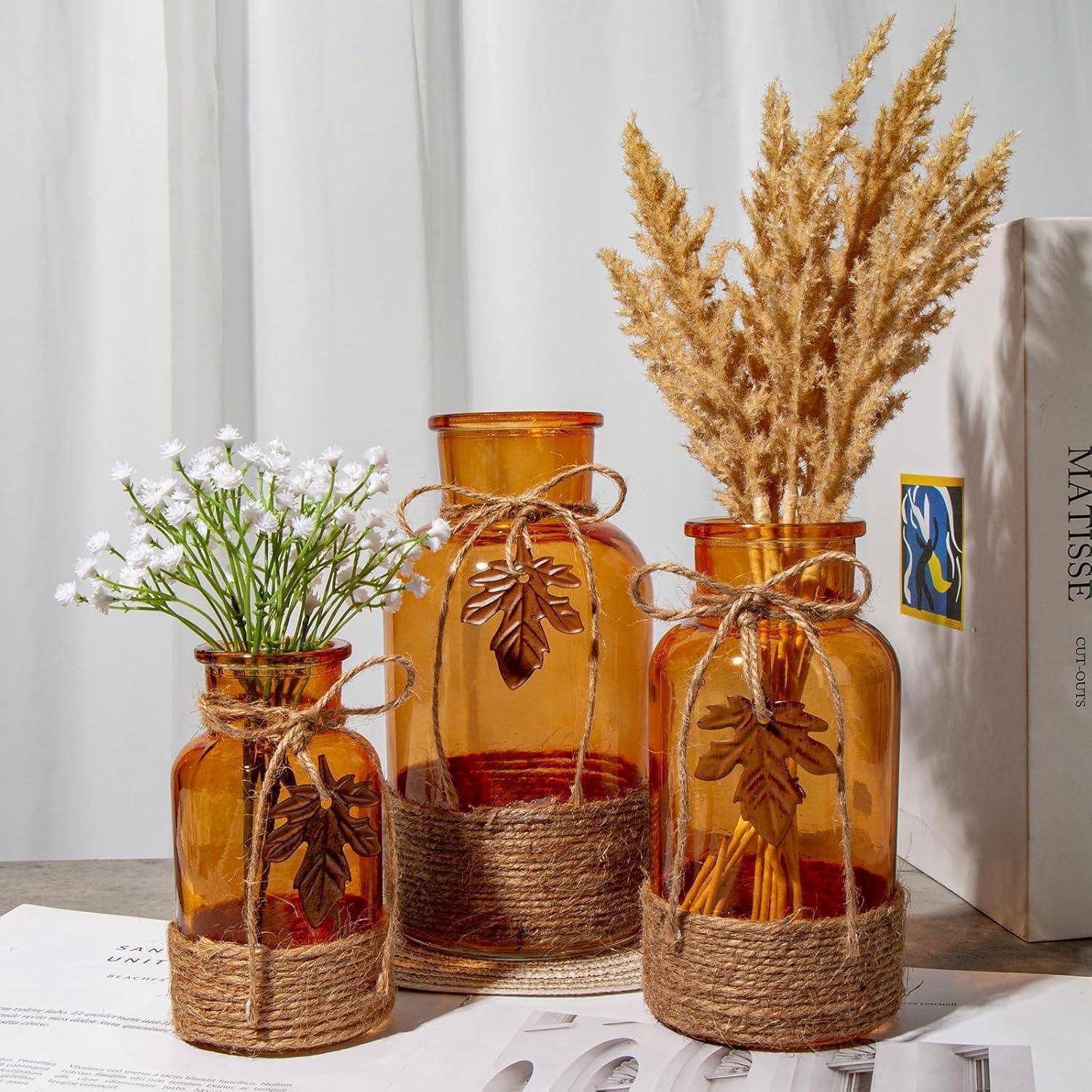 Drei Vasen aus bernsteinfarbenem Glas, mit Bindfaden umwickelt, tragen weiße Blumen und getrocknete Gräser und stehen auf einem Holztisch neben einem Stapel Bücher.