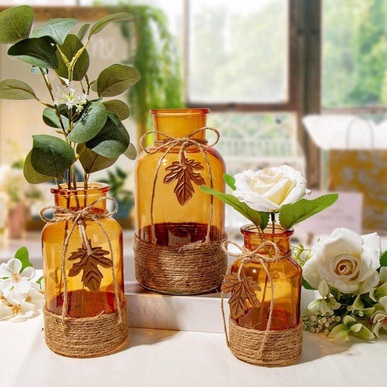 Drei Vasen aus bernsteinfarbenem Glas, mit Bindfaden umwickelt, gefüllt mit grünem Laub und einer weißen Rose, auf einem Tisch bei weichem, natürlichem Licht.
