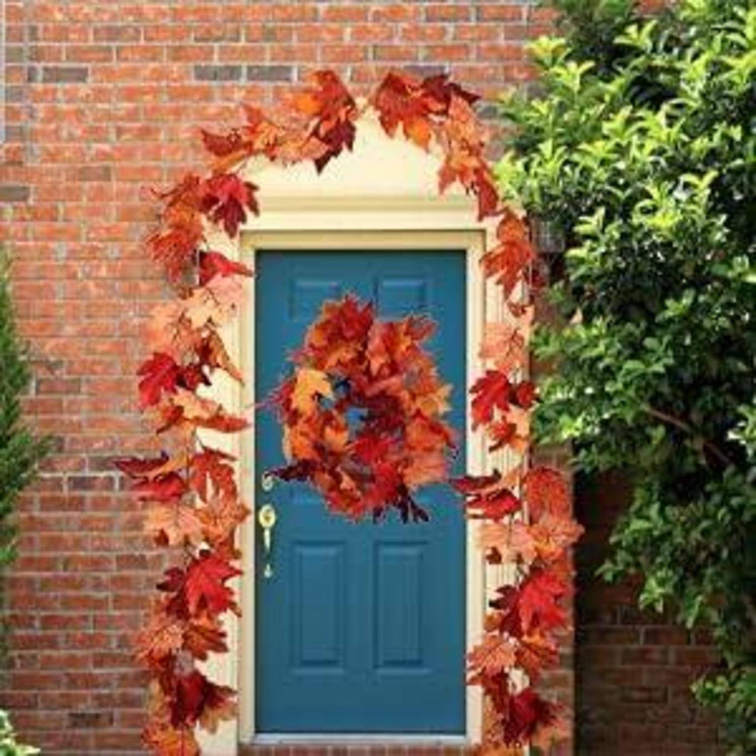 Eine blaue Tür ist mit einem Kranz aus leuchtenden Herbstblättern an einem orangefarbenen Ziegelhaus geschmückt, umrahmt von grünem Laub.