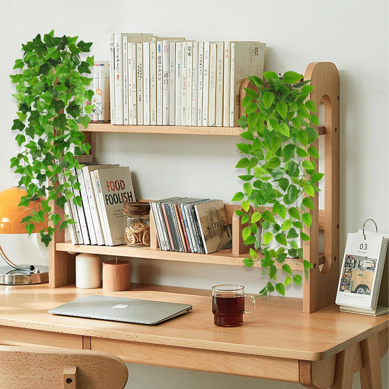 Ein aufgeräumter Holzschreibtisch mit einem Laptop, einem Teeglas, Büchern und herabhängenden grünen Pflanzen von einem Regal darüber.