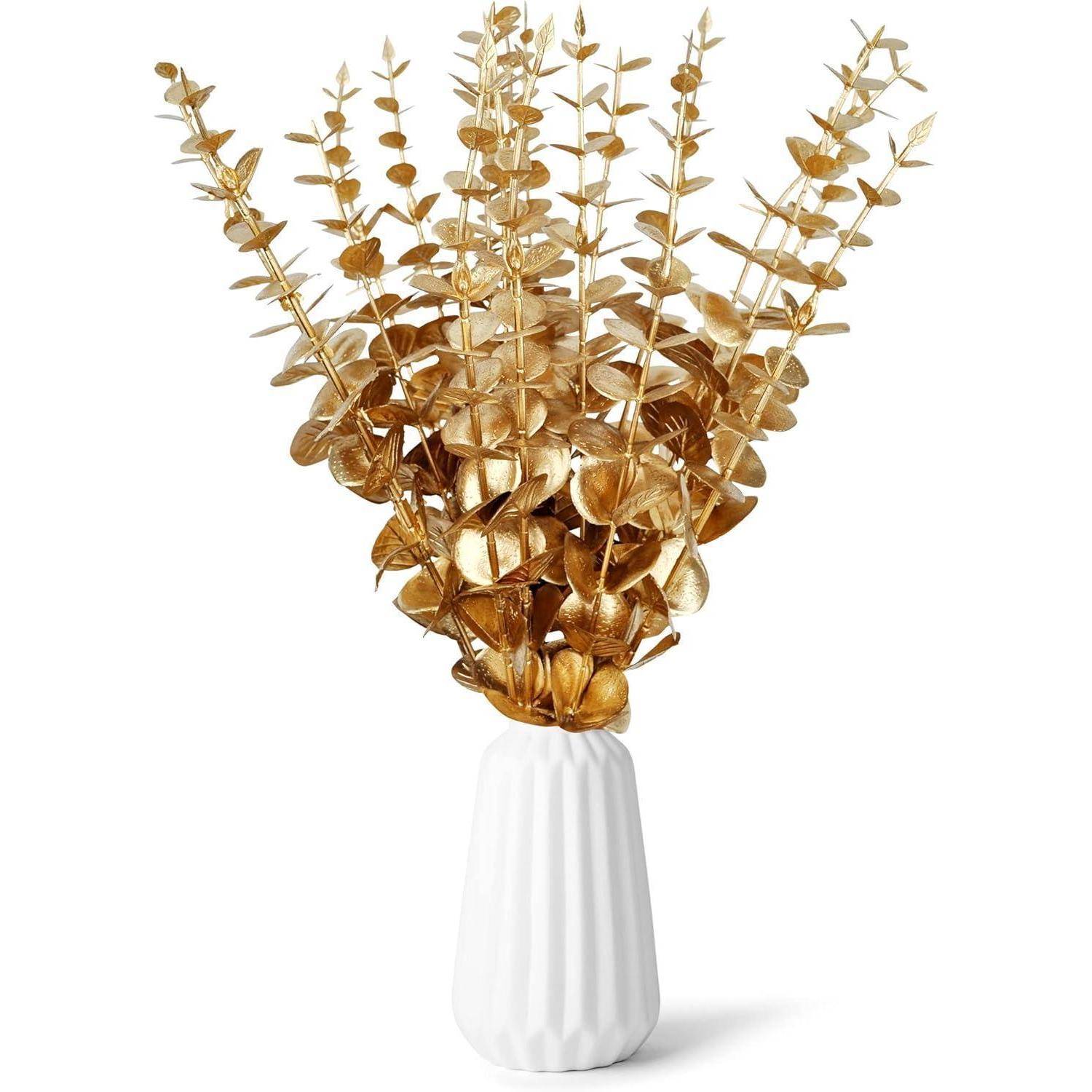 Eine weiße Vase enthält goldene Eukalyptusstiele, die sich elegant fächerförmig ausbreiten und einen luxuriösen und modernen Dekorationsarrangement schaffen.